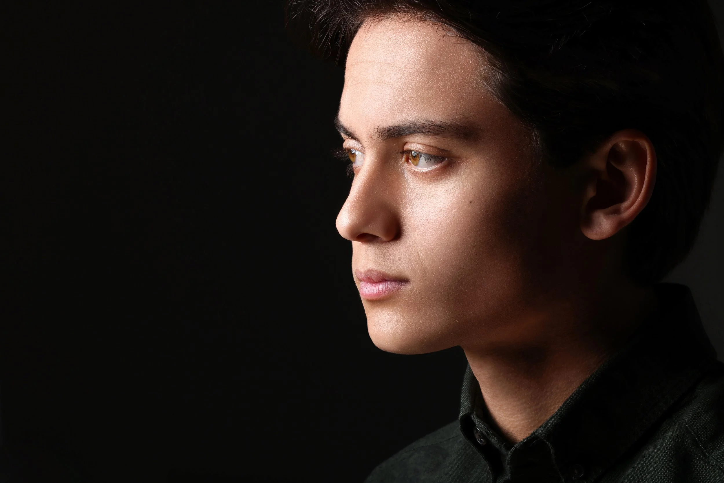 Profile of a young man with dark hair, wearing a black shirt, looking to the left against a black background.