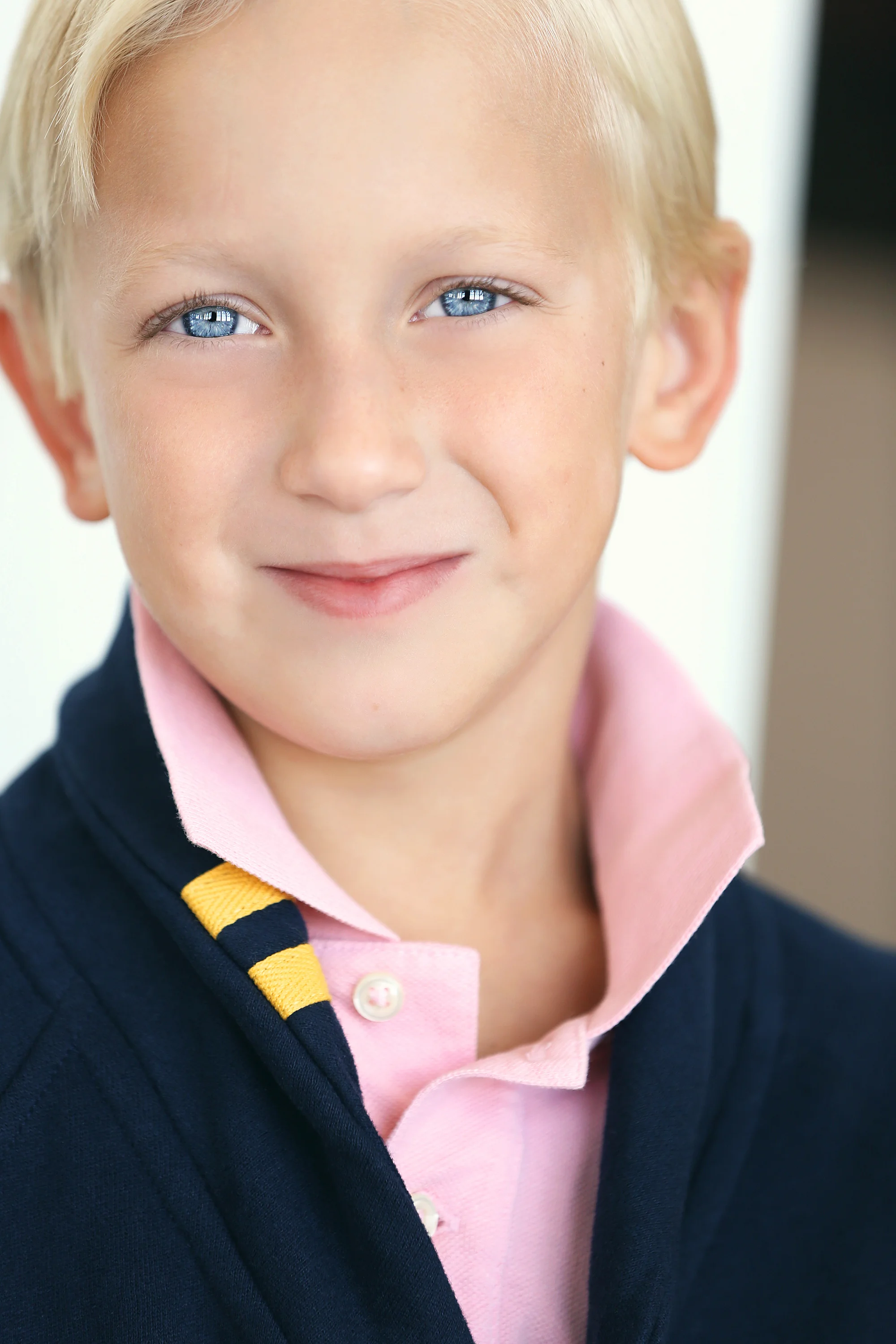 Close-up of a young boy with blonde hair and blue eyes, wearing a pink collared shirt and a navy blue sweater with yellow and black striped accents.