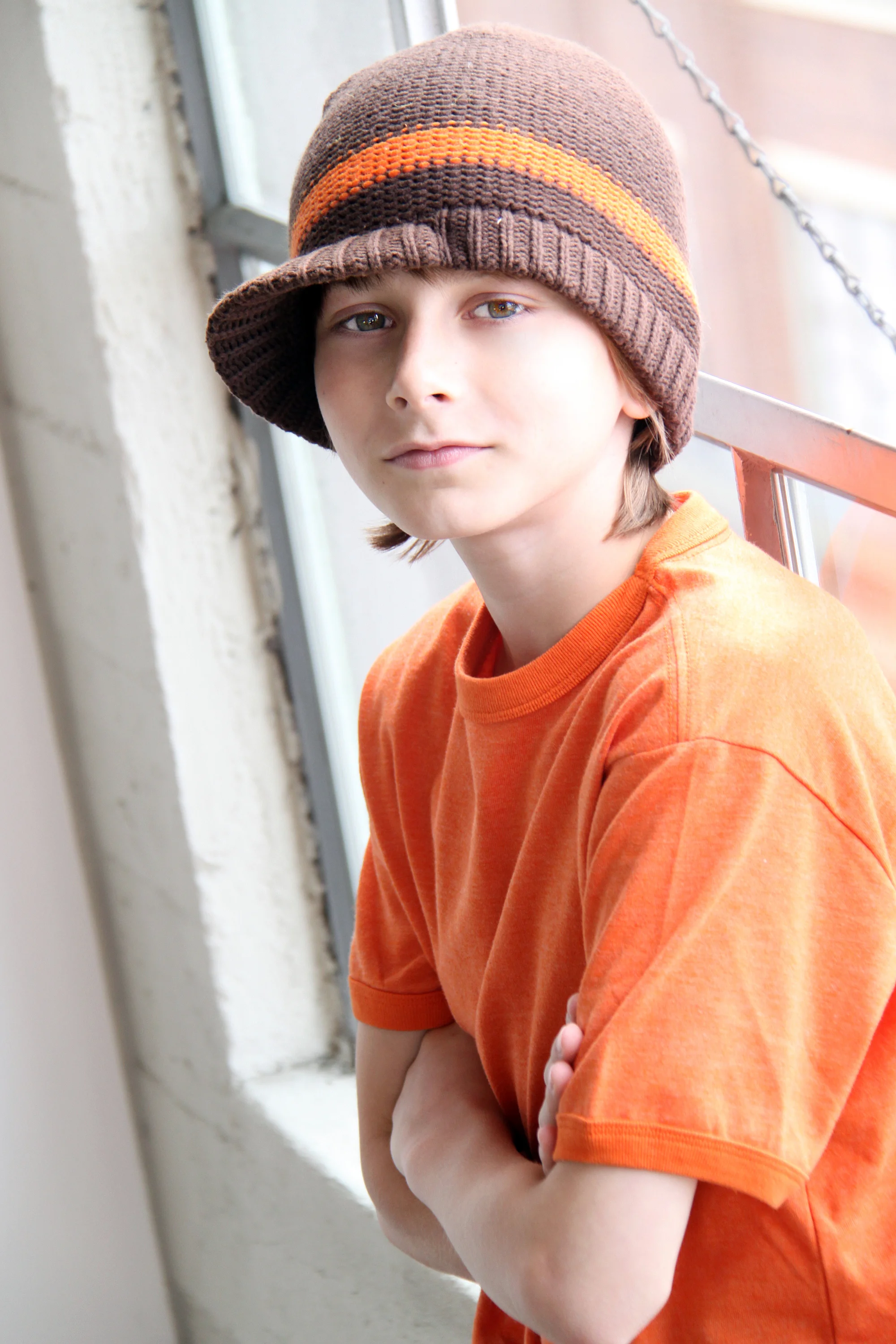 A young boy with headphones resting on his shoulders, wearing an orange t-shirt and a brown knit beanie, sitting on a windowsill.