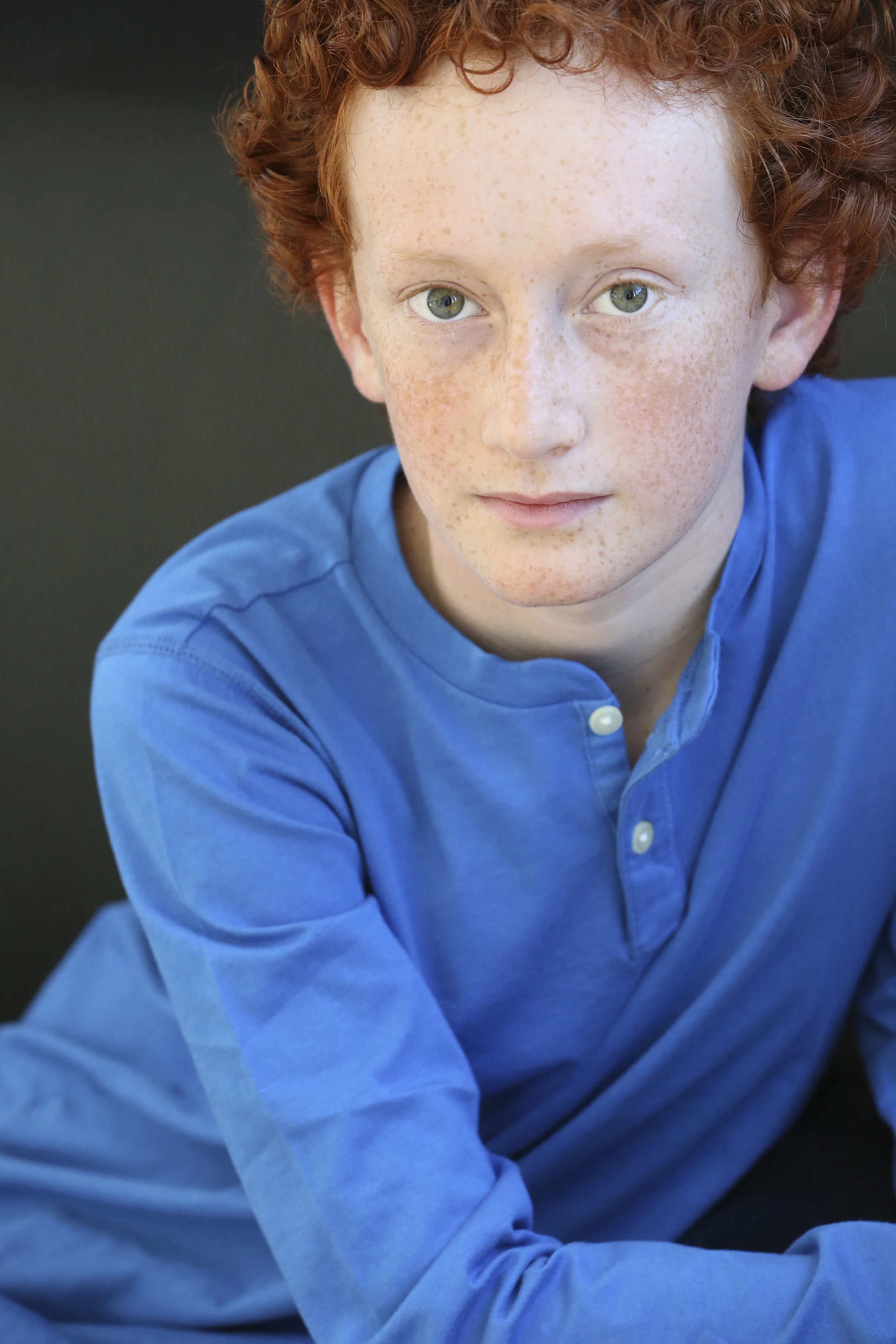 Close-up of a young boy with curly red hair and freckles, wearing a blue shirt, looking at the camera with a neutral expression.