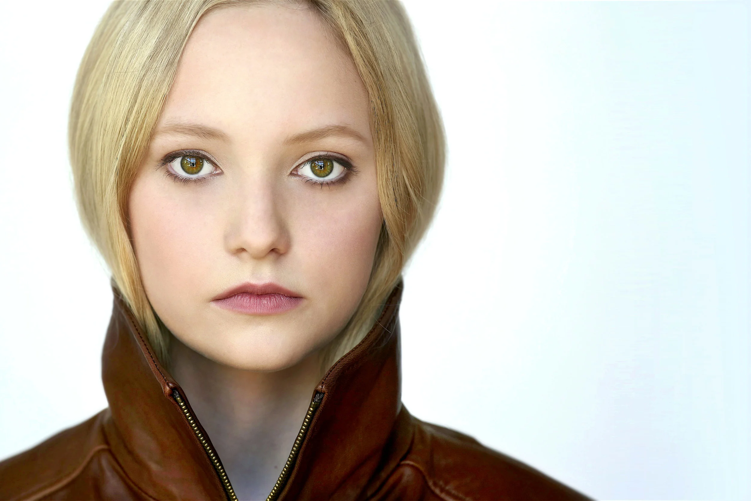 Close-up of a woman with short blonde hair, green eyes, and a serious expression, wearing a brown leather jacket with a zipper, against a plain light background.