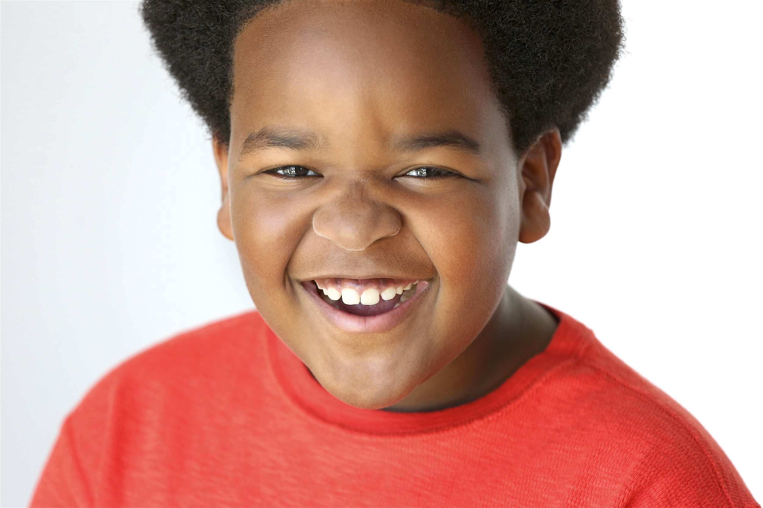 Close-up of a young boy with dark curly hair wearing a red shirt, smiling broadly with teeth showing, against a plain white background.