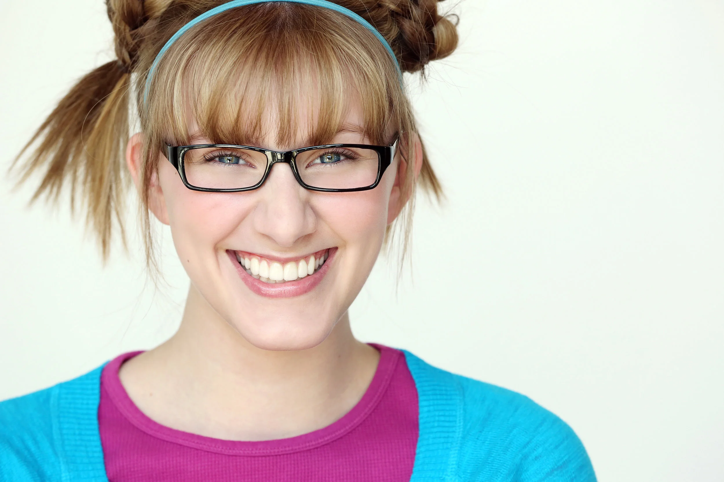 A young woman with glasses smiling, with her hair styled in pigtails and wearing a pink shirt and blue cardigan.