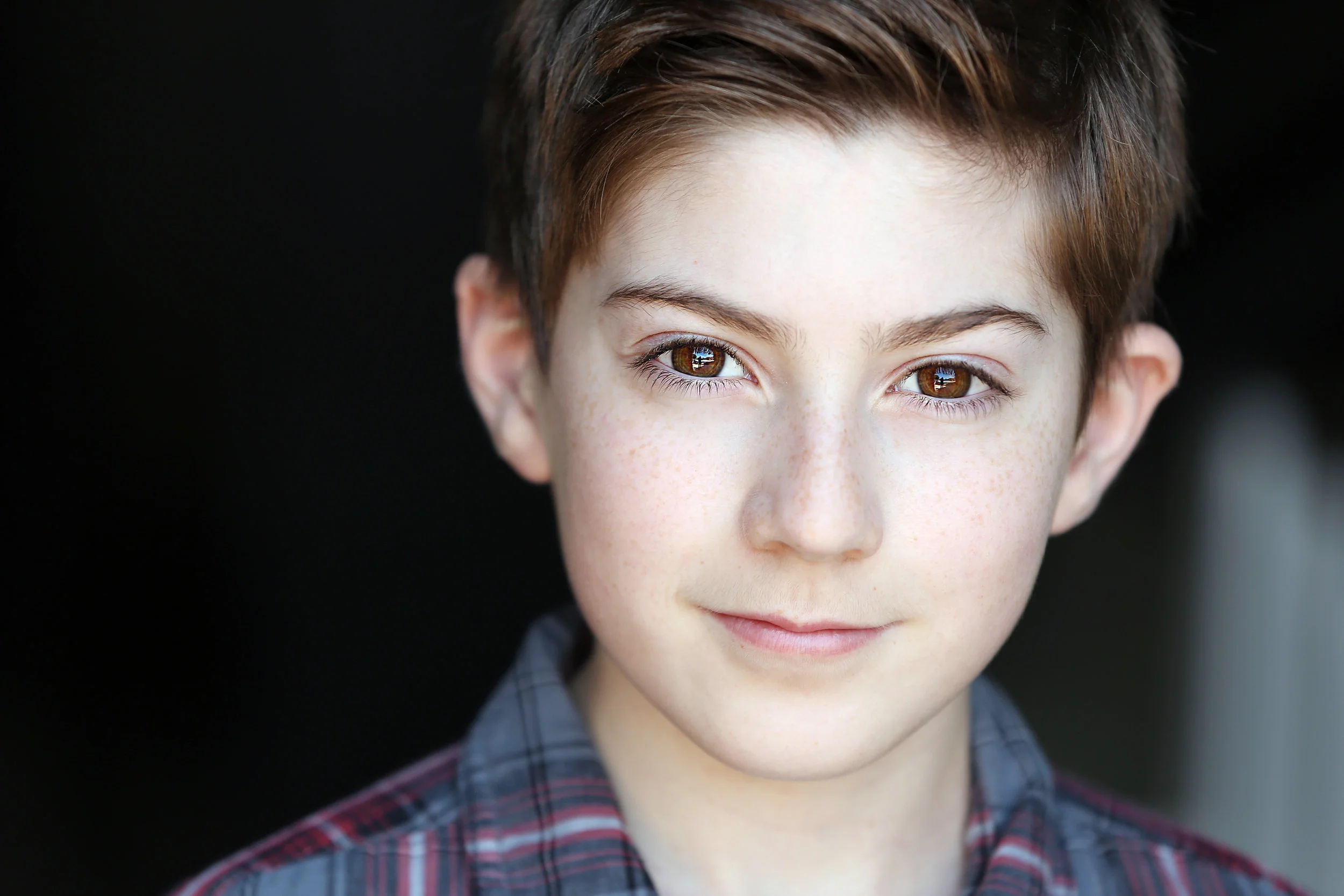Close-up of a young boy with brown hair and hazel eyes, smiling softly, wearing a plaid shirt.