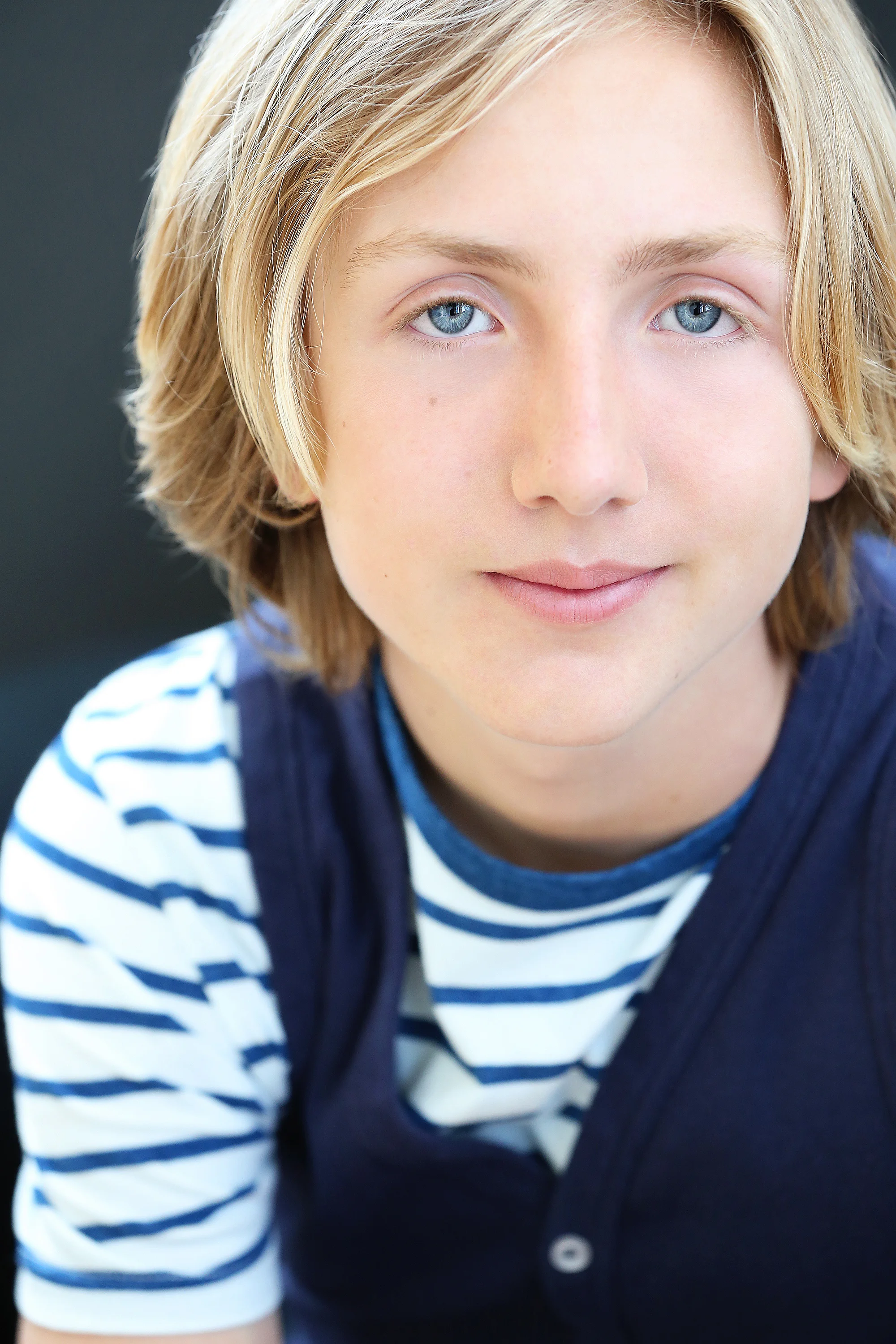 Close-up of a young boy with blond hair and blue eyes, wearing a blue and white striped shirt and navy vest, smiling softly.