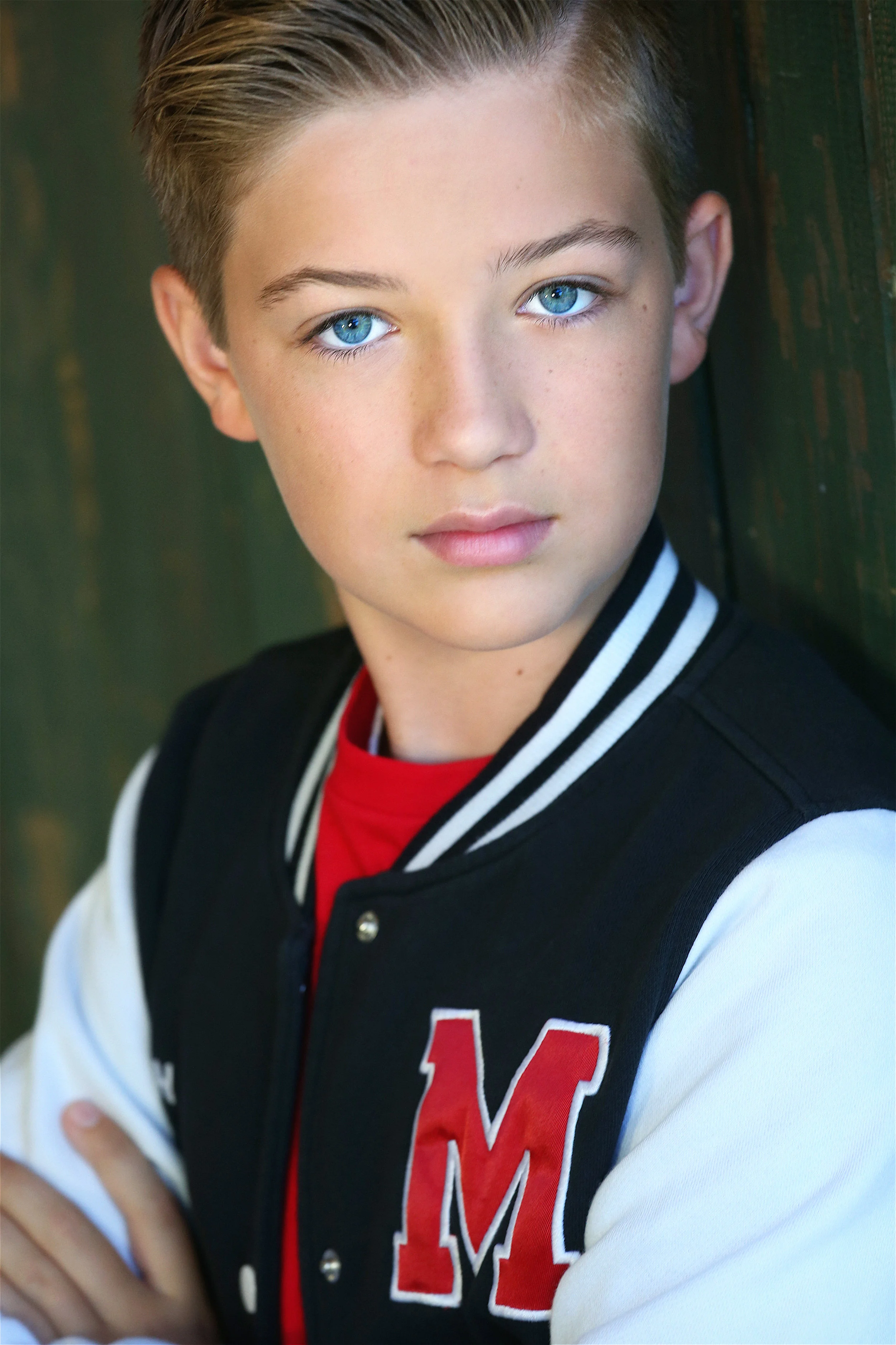 Close-up of a teenage boy with blue eyes and short light brown hair, wearing a varsity jacket with a red letter M on it, leaning against a wooden green wall.
