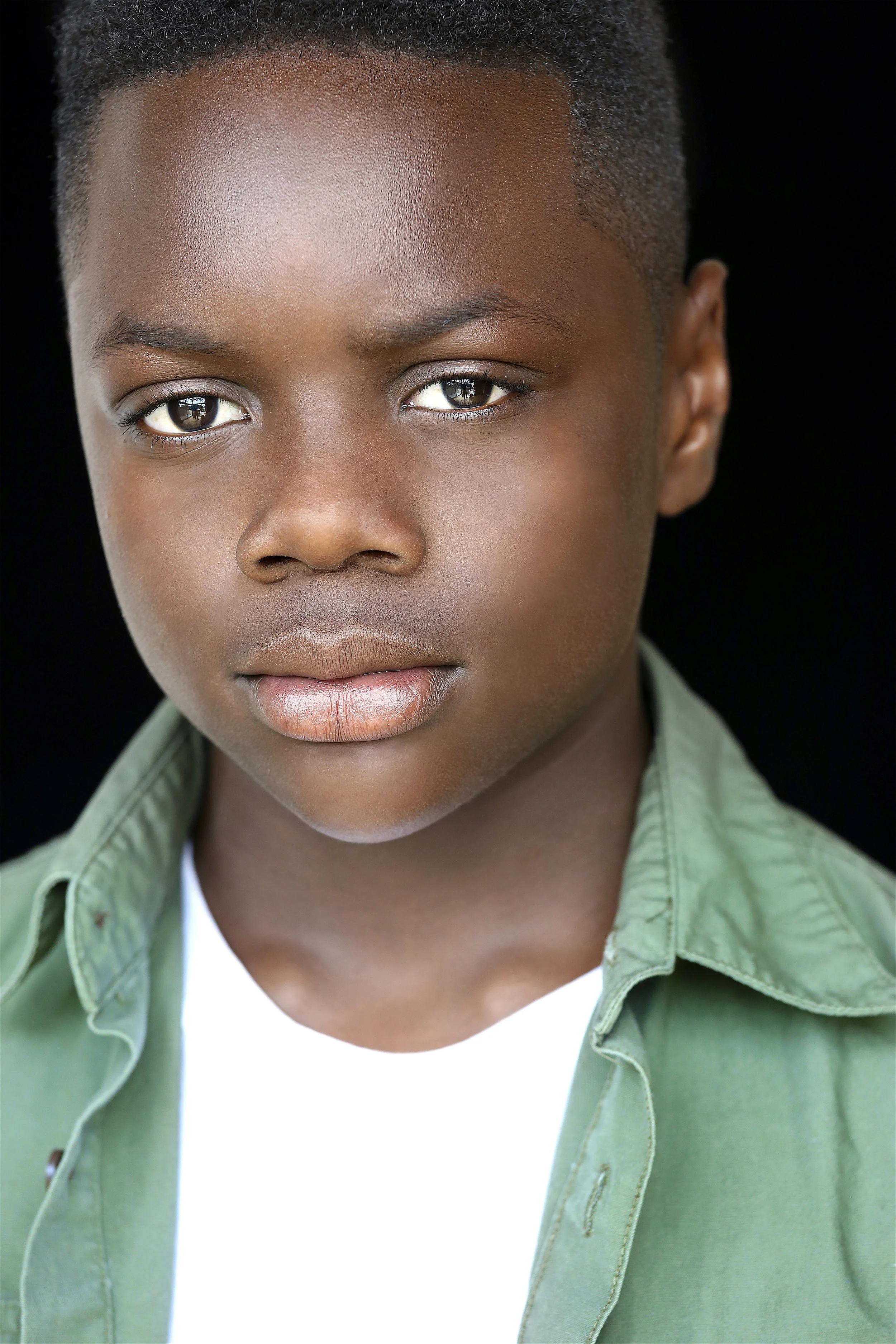 Close-up portrait of a young boy with dark skin and short curly hair, wearing a green button-up shirt over a white T-shirt, against a black background.