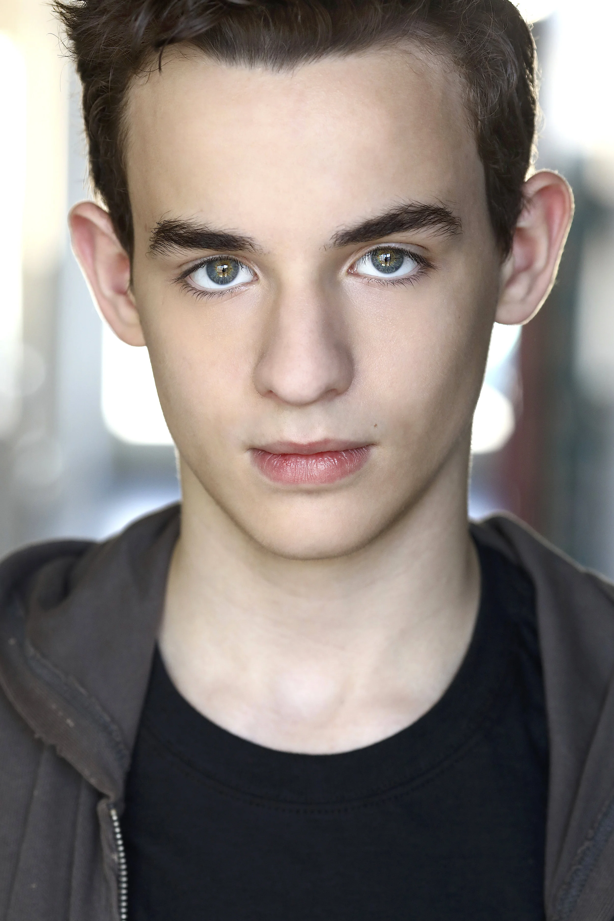 Close-up of a young man with striking blue eyes, dark hair, and fair skin, wearing a black shirt and dark jacket, indoors with a blurred background.