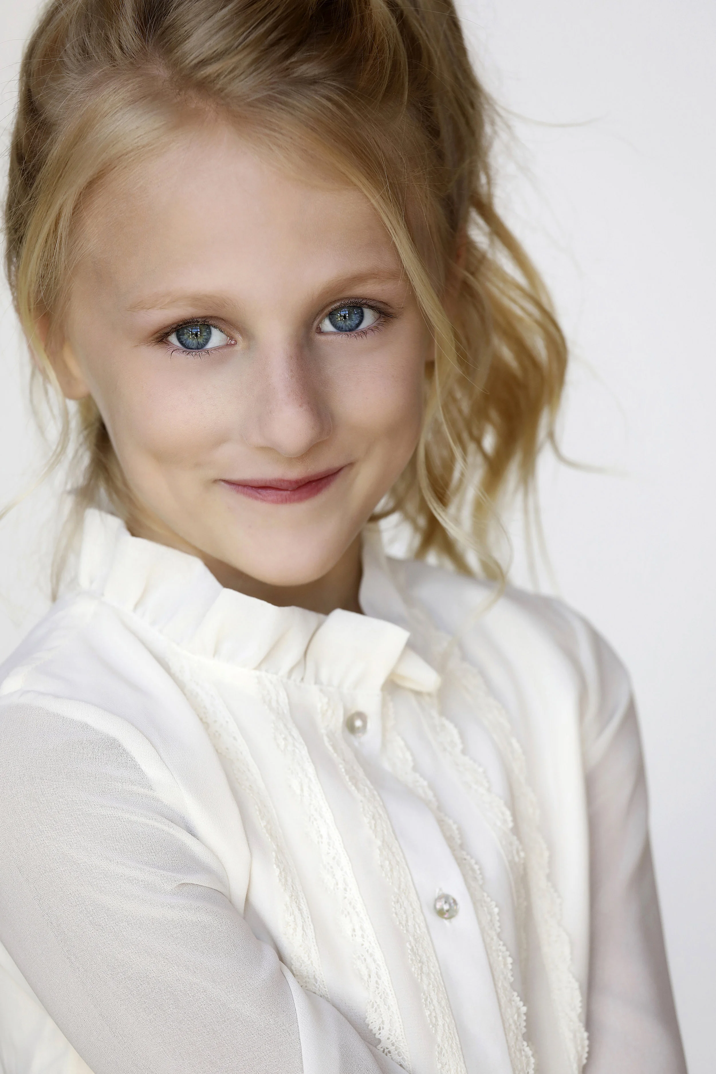 Close-up of a young girl with blonde hair and blue eyes wearing a white blouse smiling softly.