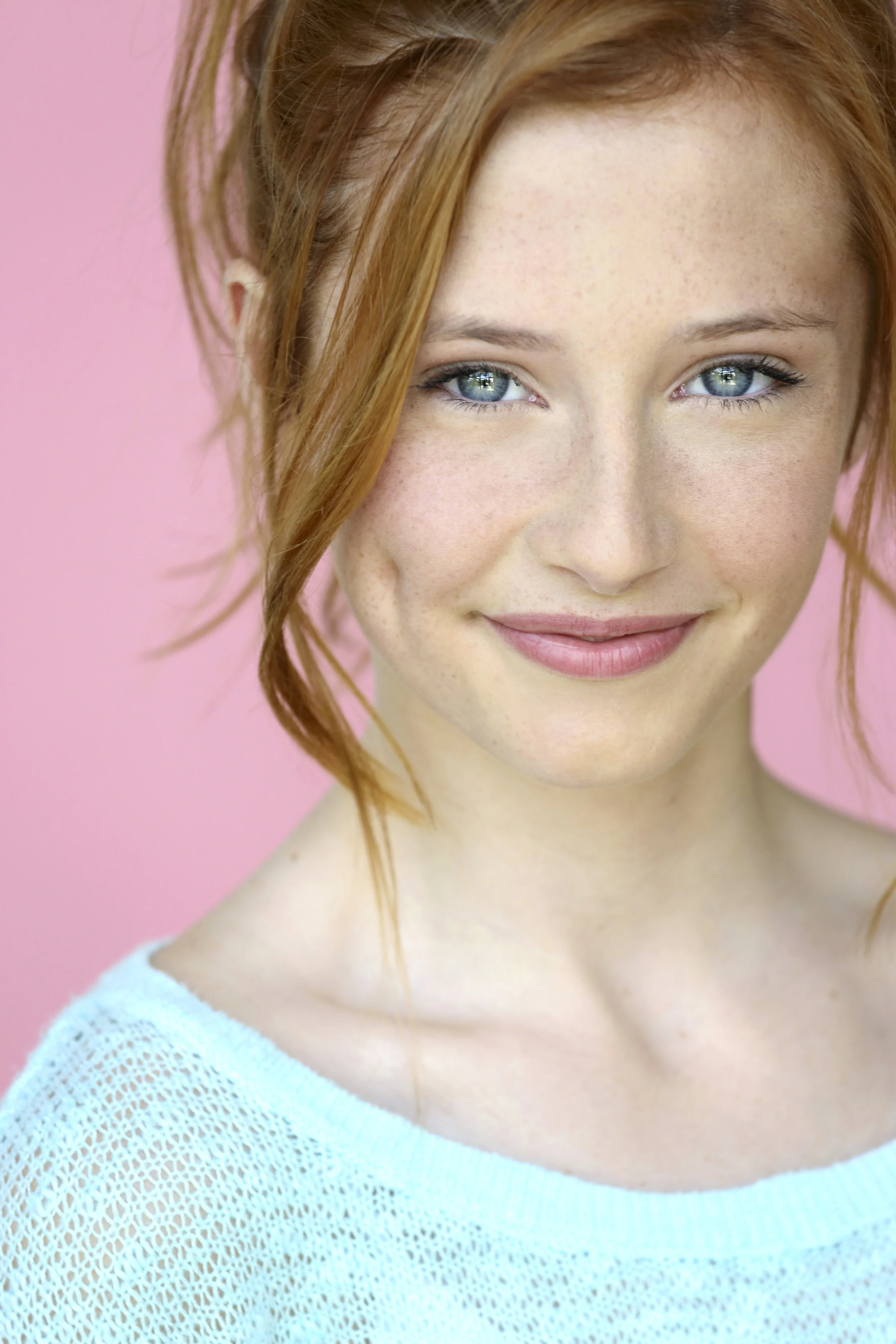 Close-up portrait of a young woman with red hair, blue eyes, and fair skin with freckles, smiling against a pink background.
