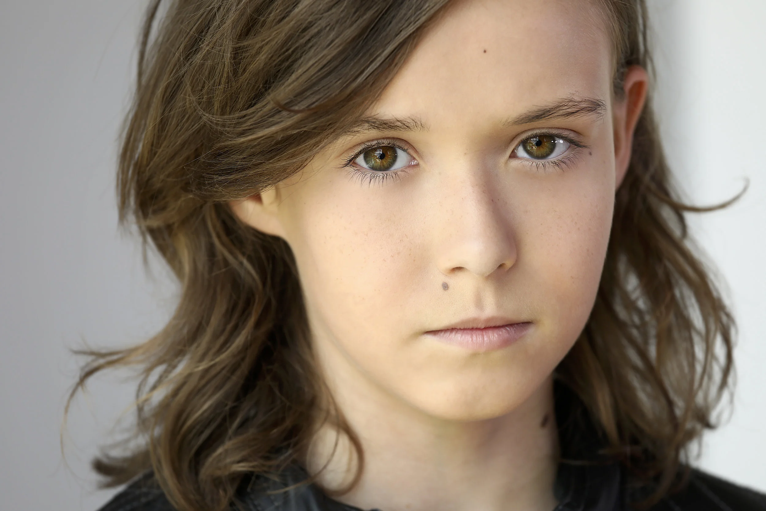 Close-up of a young girl with light brown hair, hazel eyes, and a mole on her cheek, looking directly at the camera.