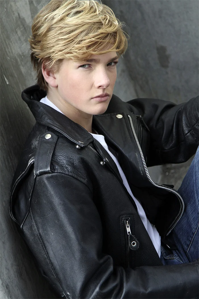 Young person with blonde hair wearing a black leather jacket, sitting against a concrete wall, looking serious or contemplative.