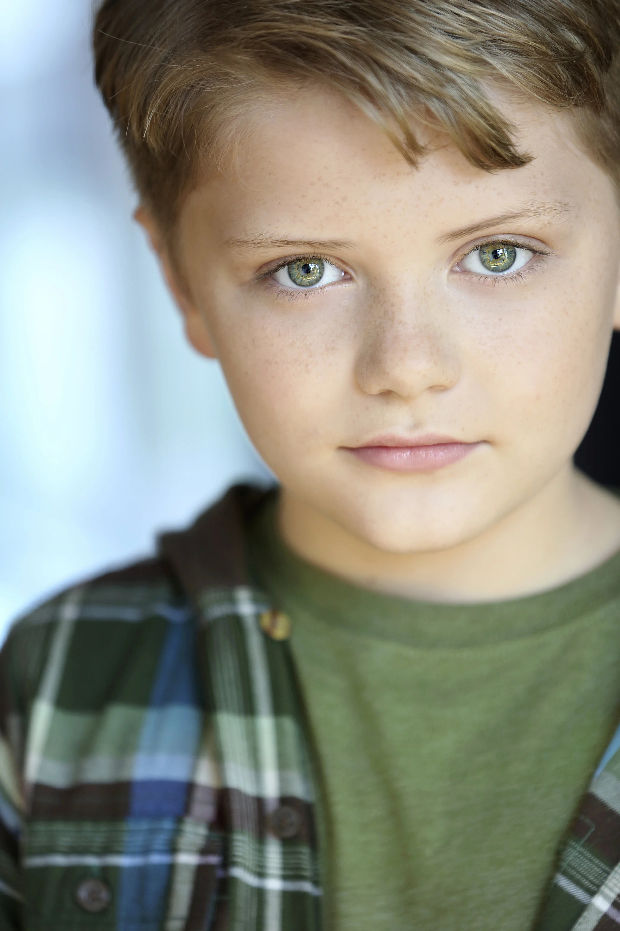 Close-up portrait of a young boy with green eyes, light brown hair, freckles, wearing a green shirt and a plaid jacket.