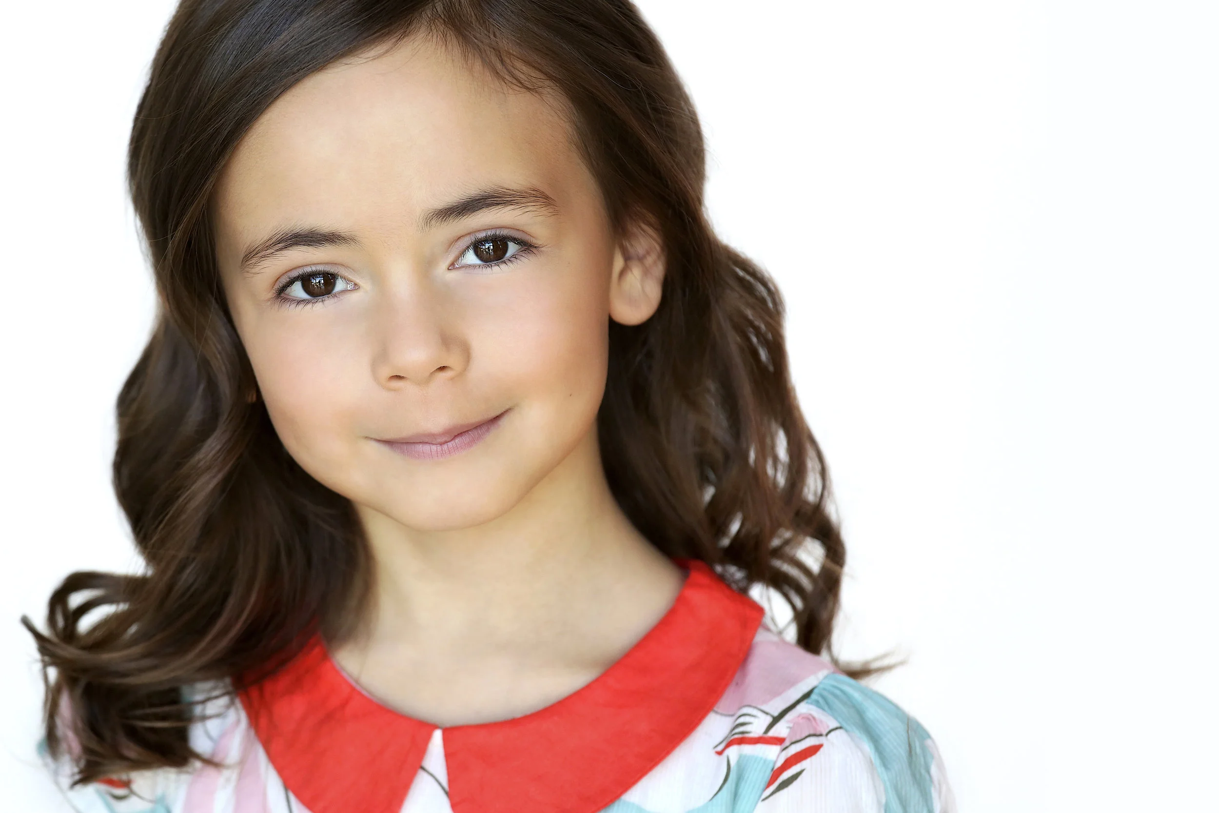 A young girl with brown hair and brown eyes, wearing a colorful top with a red collar, smiling softly against a white background.