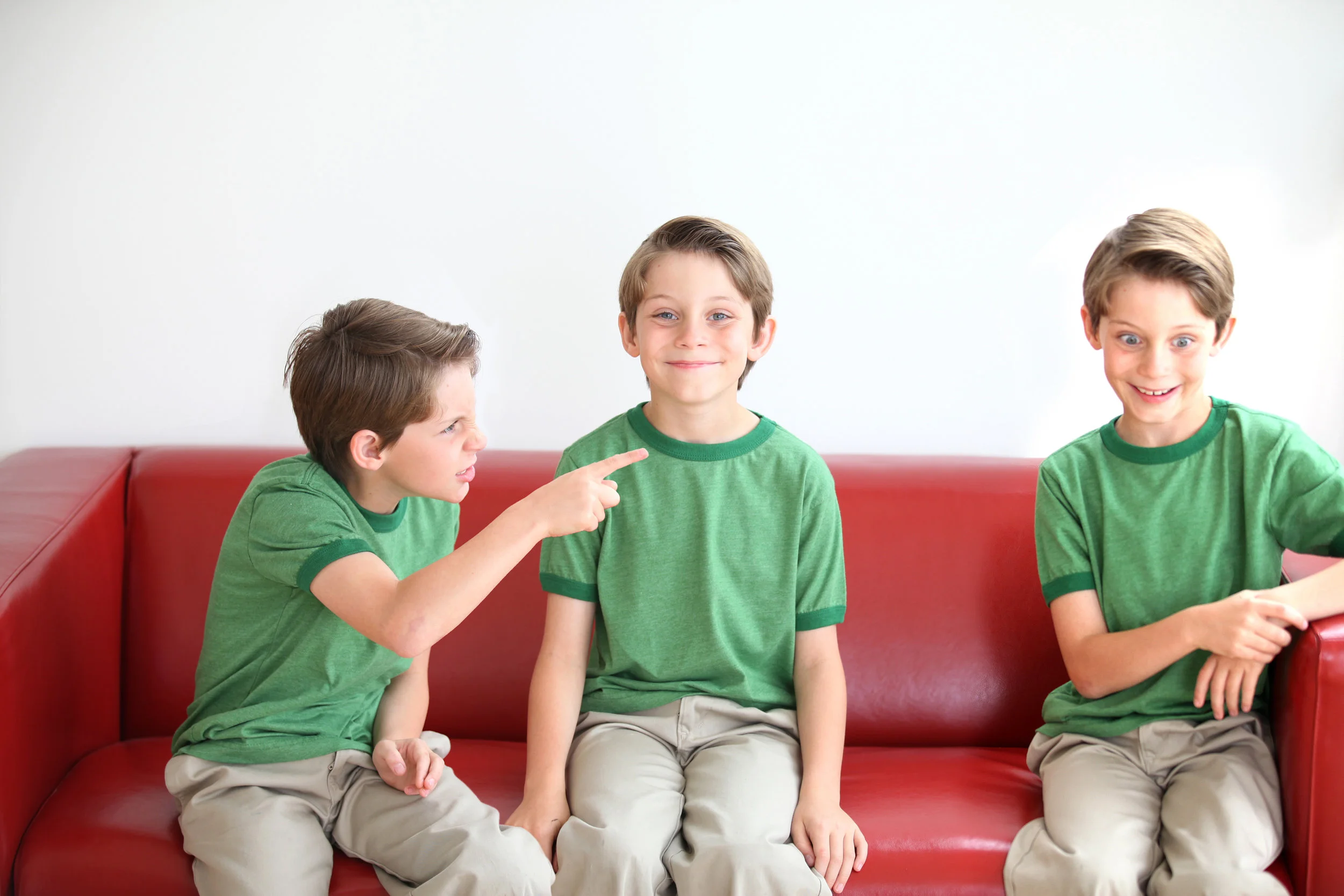 Three young boys sitting on a red couch, wearing matching green shirts and beige pants, with the boy on the left pointing and speaking to the boy in the middle, who has a slight smile, while the boy on the right looks surprised or excited.