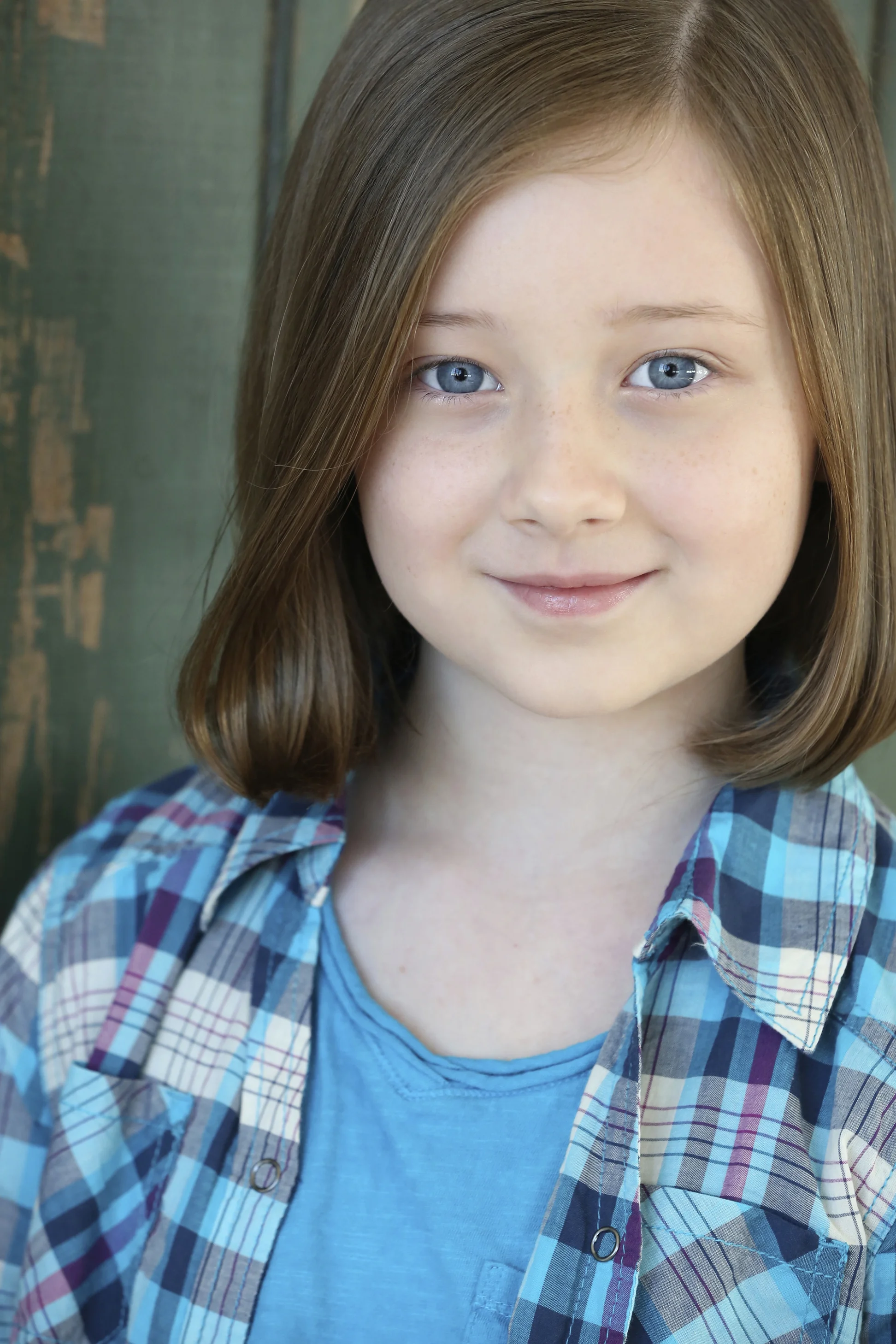 Young girl with blue eyes and shoulder-length brown hair smiling, wearing a blue shirt and plaid shirt, standing in front of a wooden background.