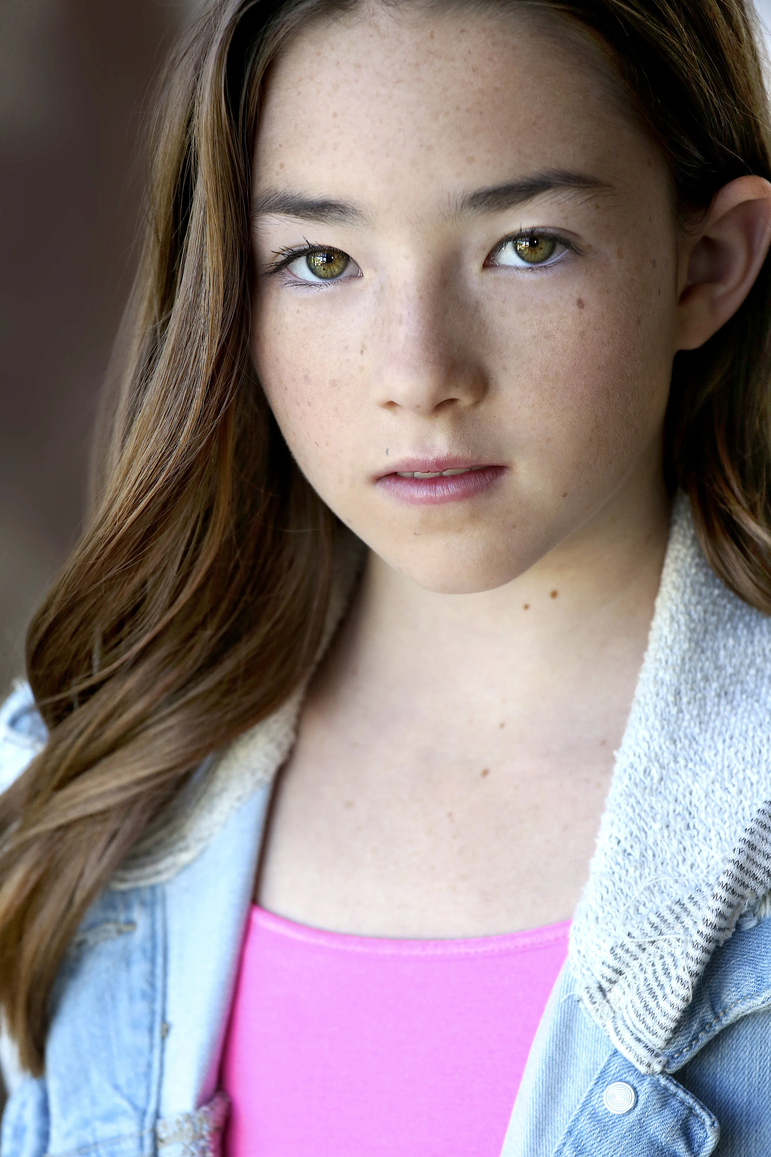 Close-up portrait of a young woman with long brown hair, green eyes, and light freckles, wearing a denim jacket and pink top.