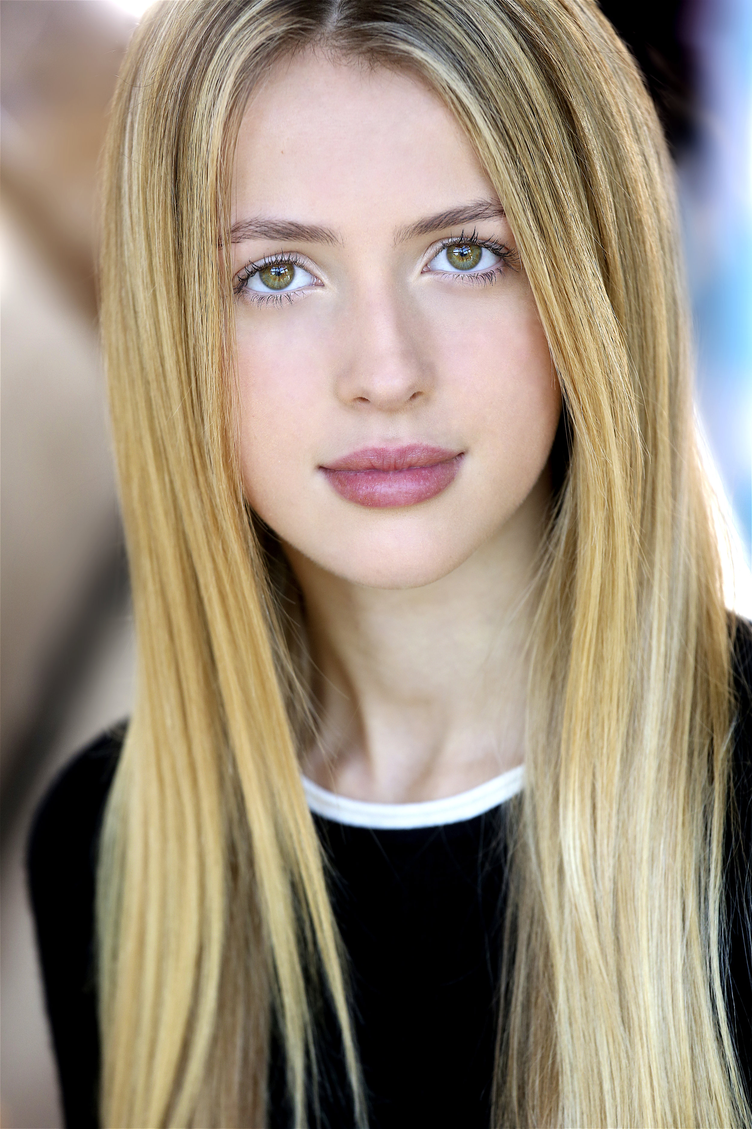 Close-up portrait of a young woman with long blonde hair, light skin, and hazel eyes, wearing a black top with a white collar.