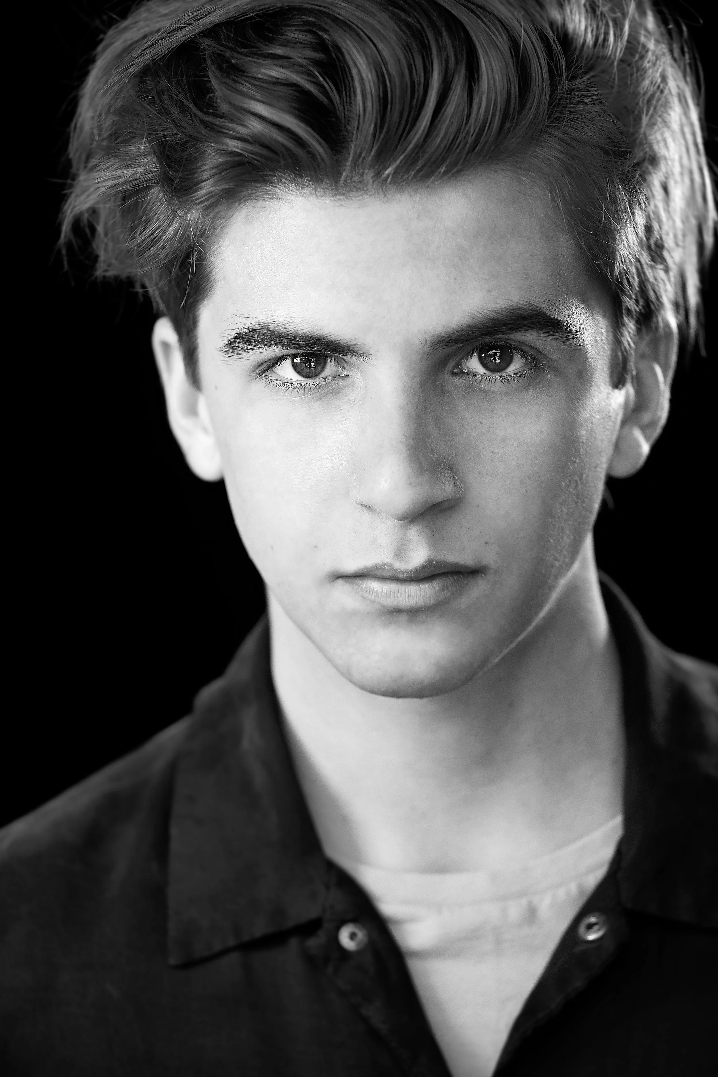 Black and white portrait of a young man with styled hair, intense gaze, wearing a collared shirt and a light-colored T-shirt underneath.