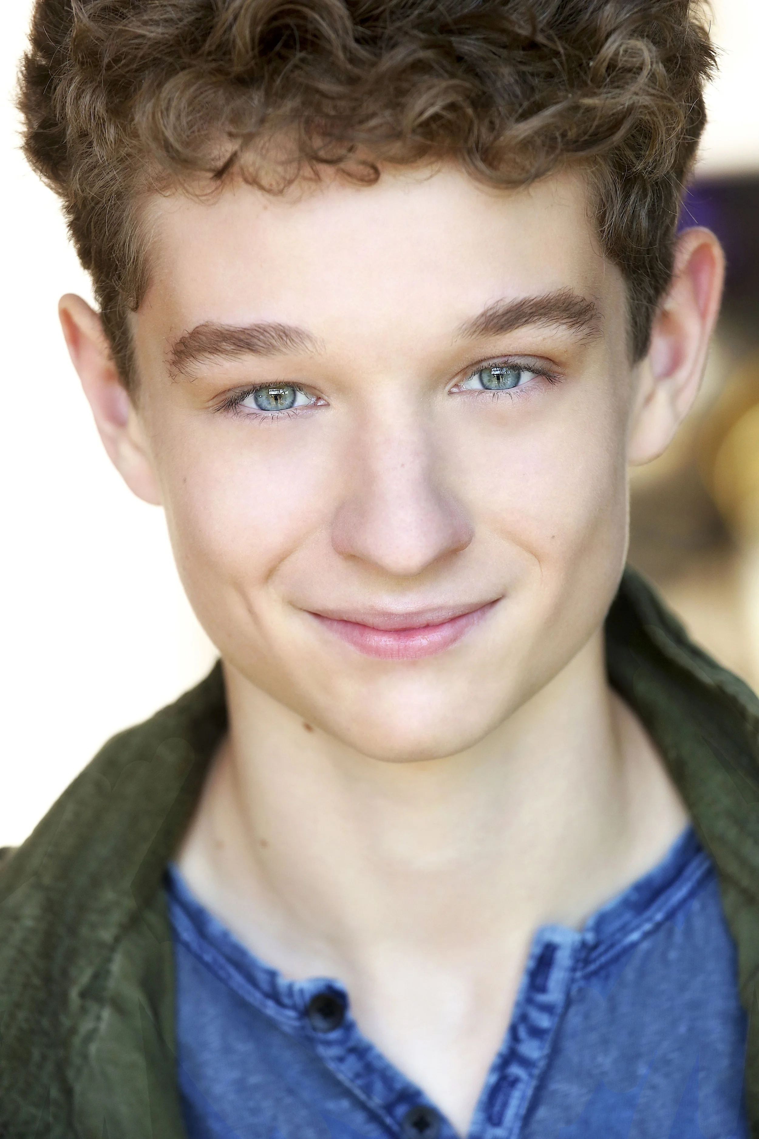 Close-up of a smiling young man with curly brown hair and blue eyes, wearing a blue shirt and green jacket.