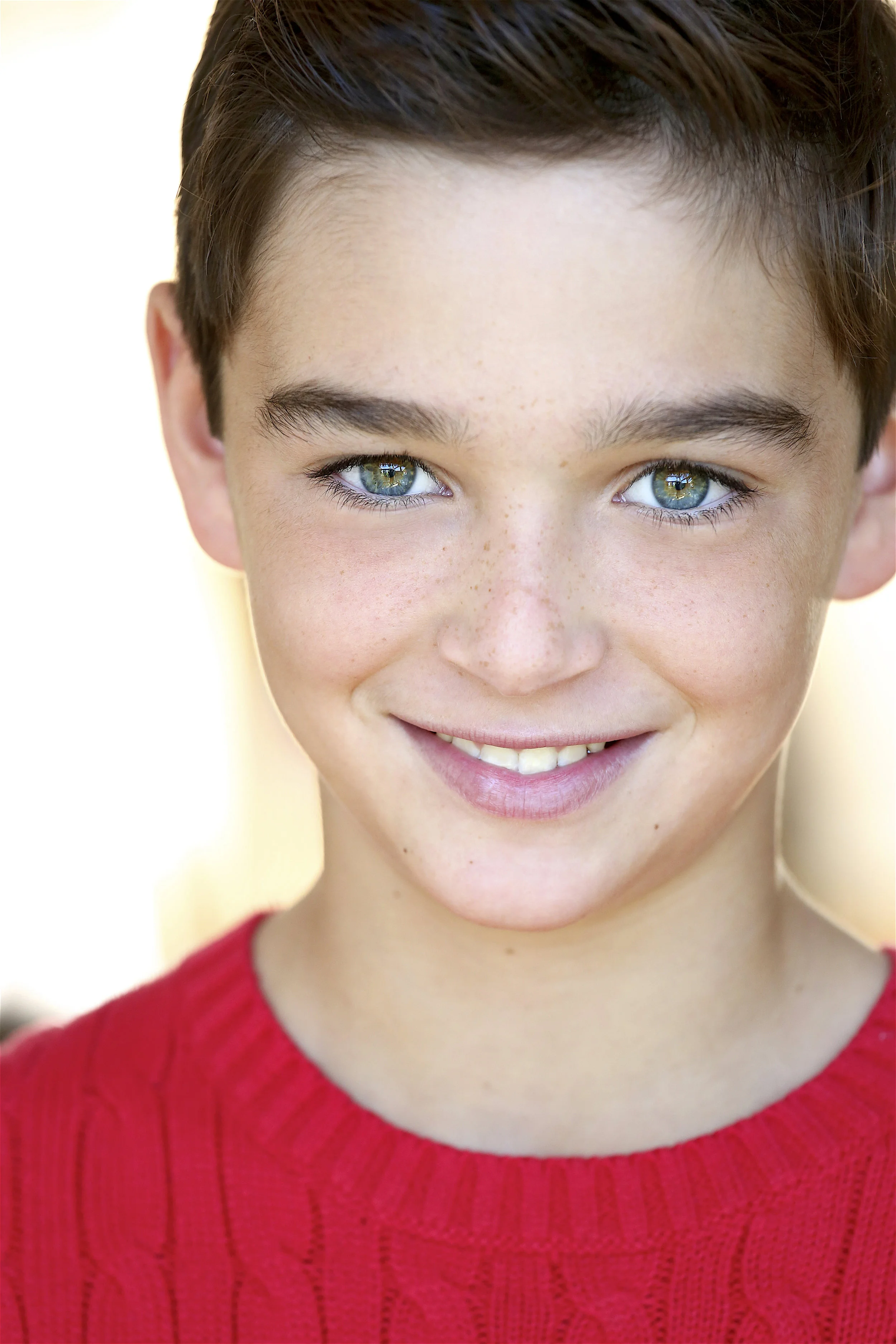 Close-up of a smiling young boy with blue eyes, freckles, and brown hair, wearing a red sweater.