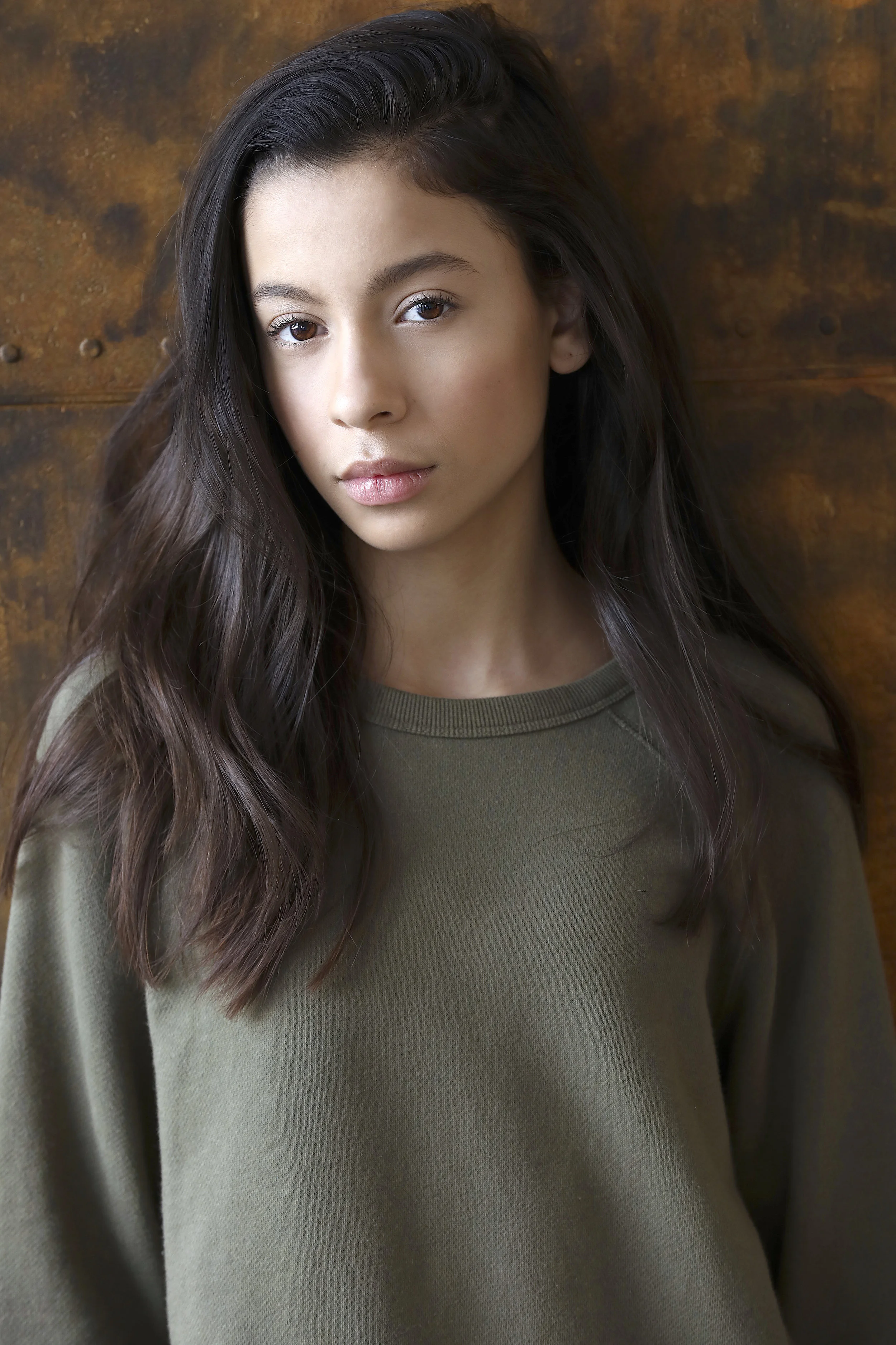Portrait of a young woman with long, wavy dark hair and light skin, wearing a green sweatshirt, standing against a wooden background.