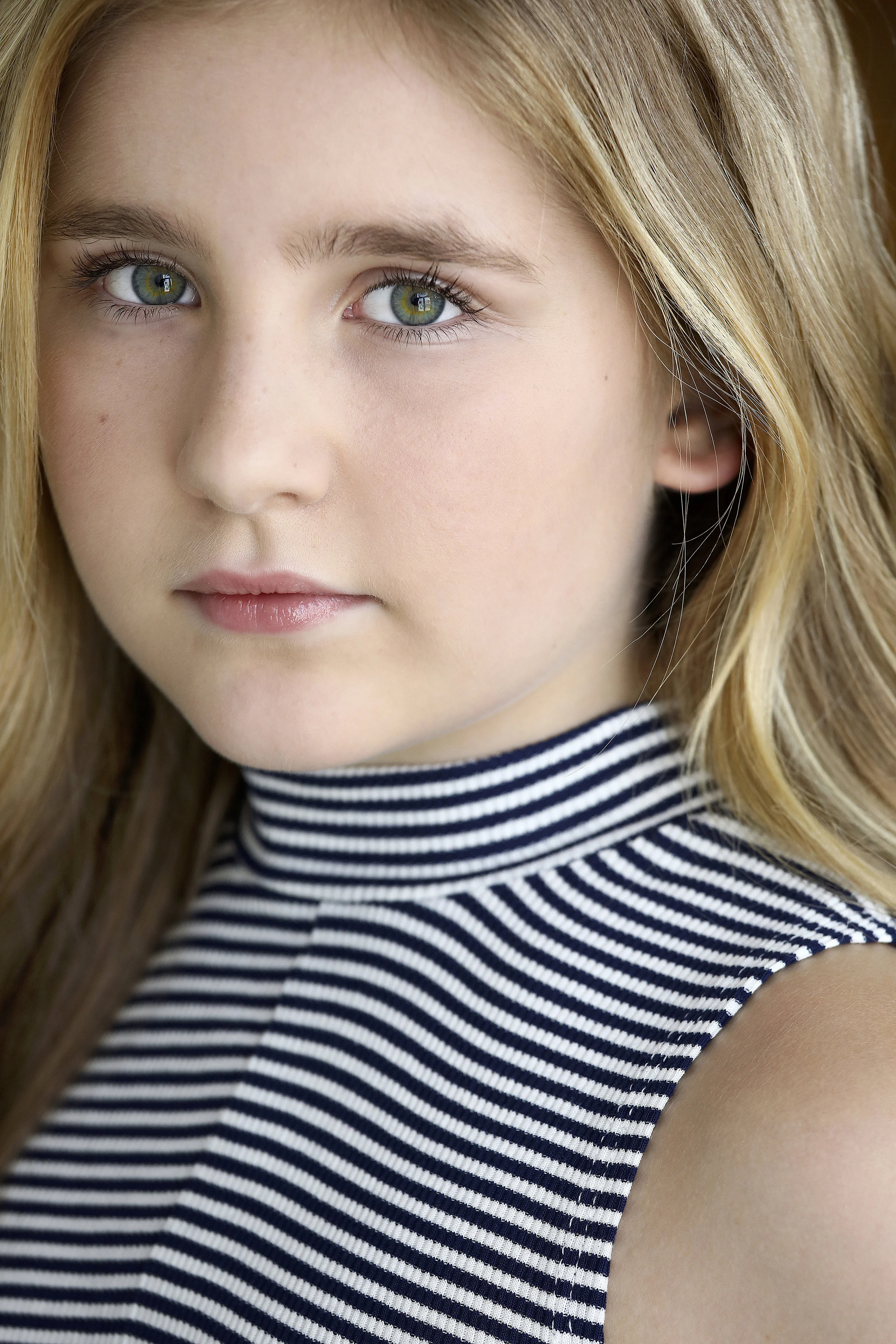 Close-up portrait of a young girl with blonde hair, green eyes, and light skin, wearing a black and white striped sleeveless turtleneck.
