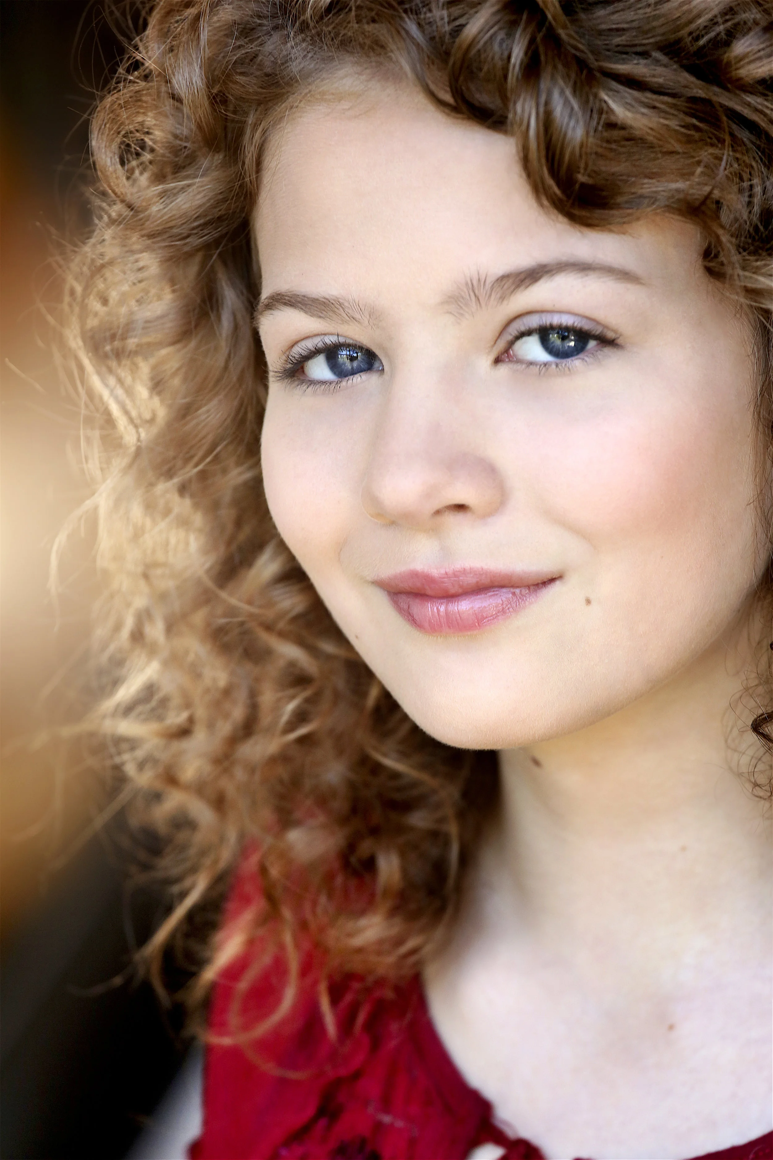 Close-up portrait of a young woman with curly red hair, blue eyes, and fair skin, wearing a red top with lace details.