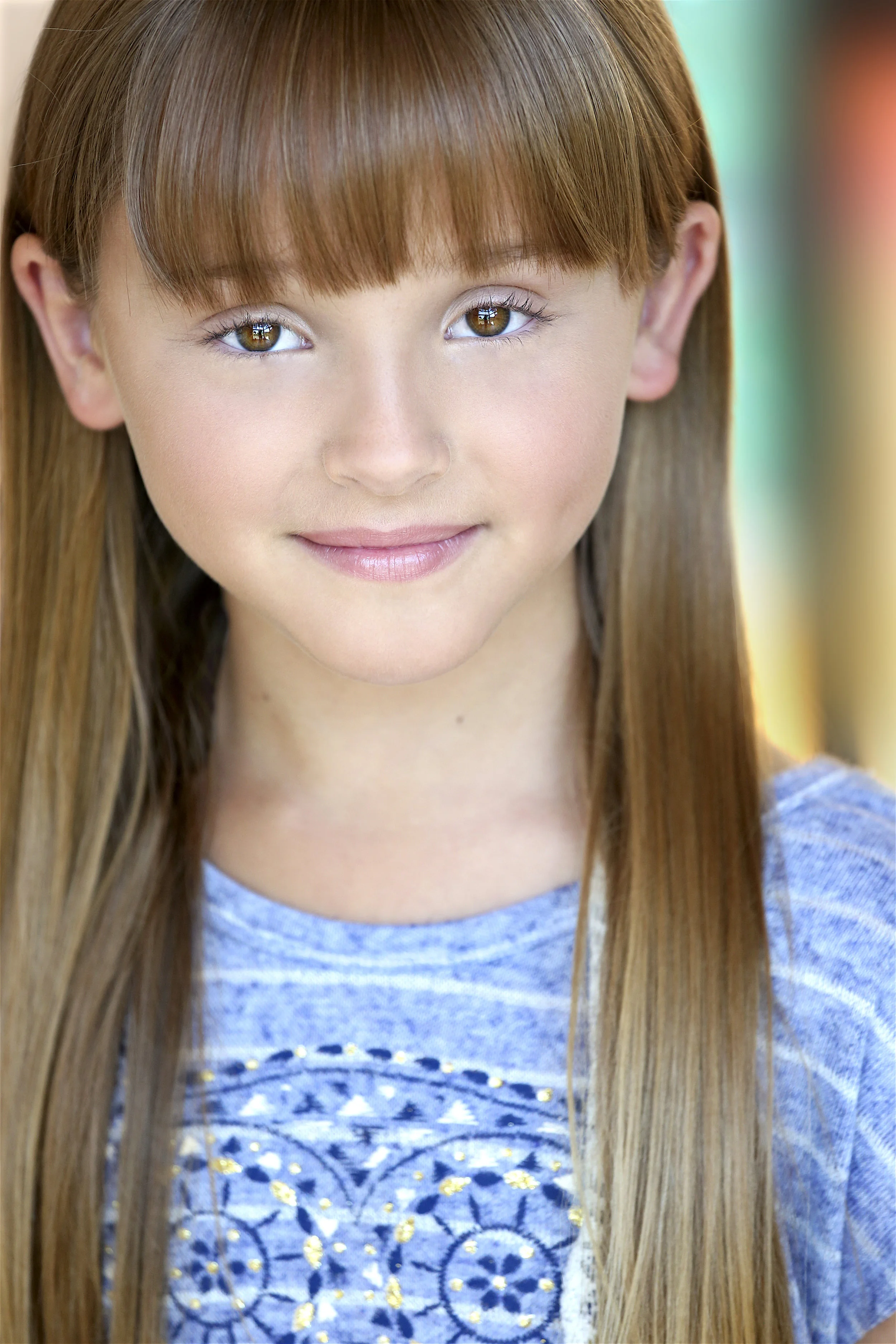 Close-up portrait of a young girl with long brown hair and brown eyes, smiling softly, wearing a blue top with embroidery, with a blurred colorful background.
