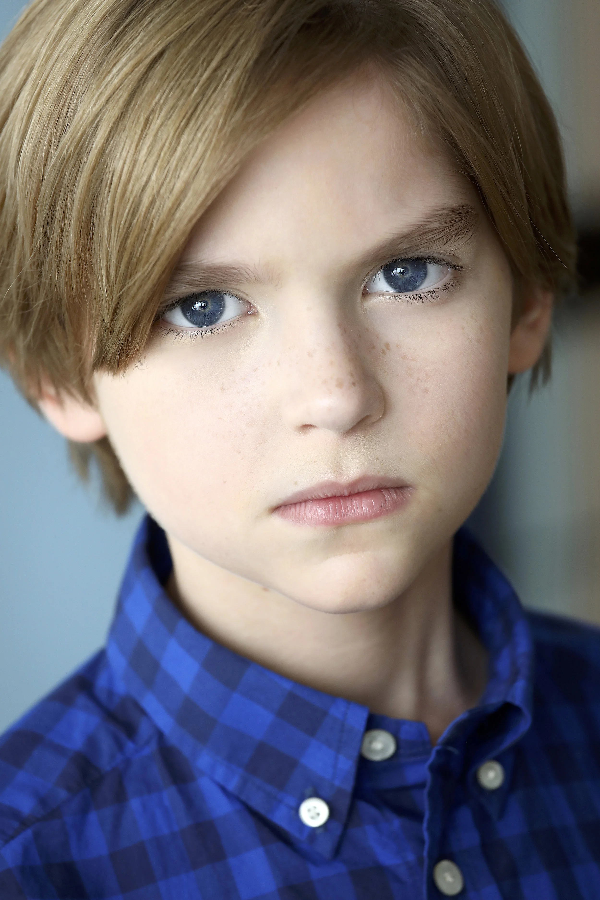 Close-up portrait of a young boy with blue eyes, light brown hair, and freckles, wearing a blue checkered shirt.