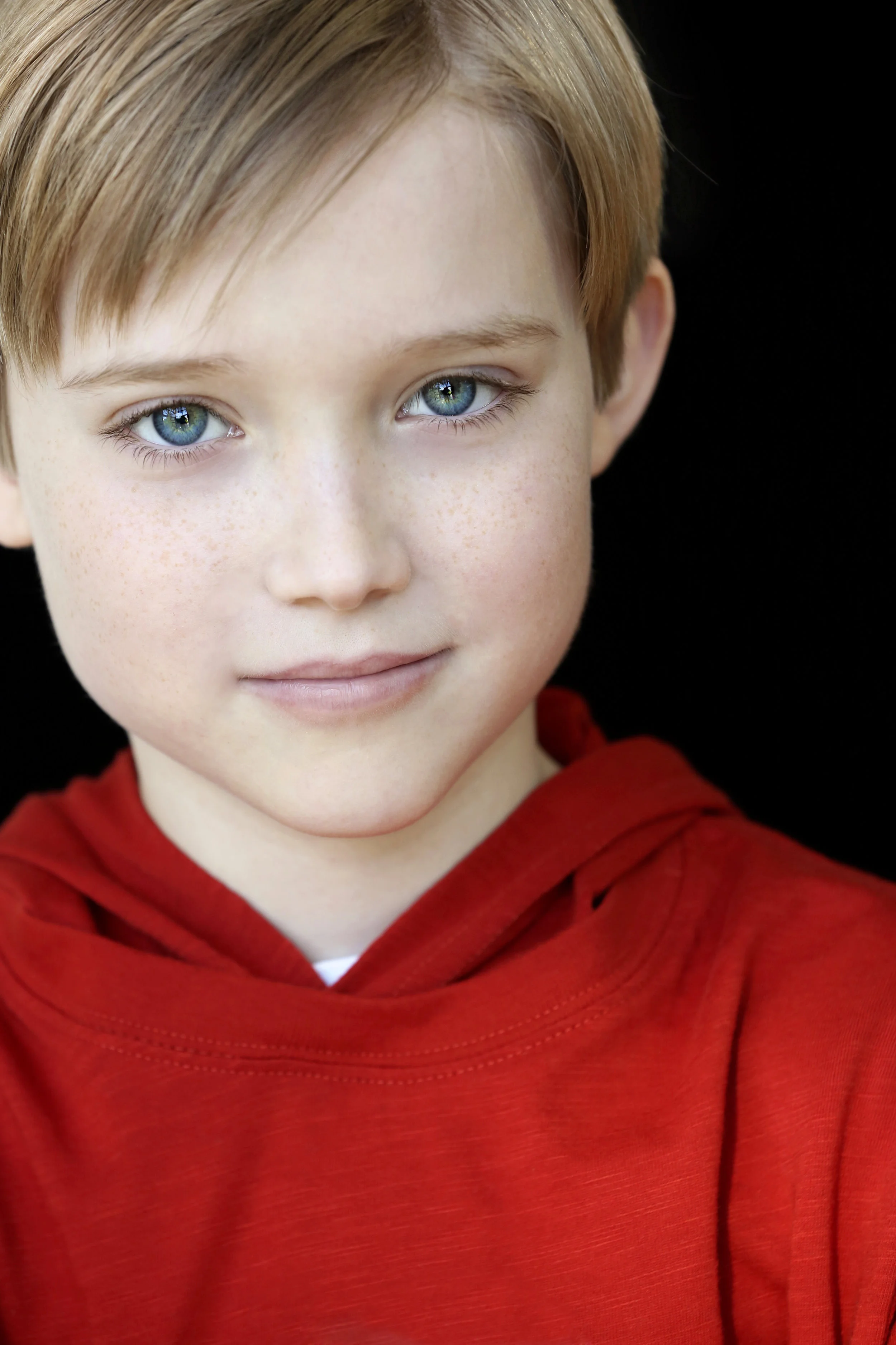 Close-up portrait of a young boy with blue eyes, freckles, and light brown hair wearing a red hoodie against a black background.