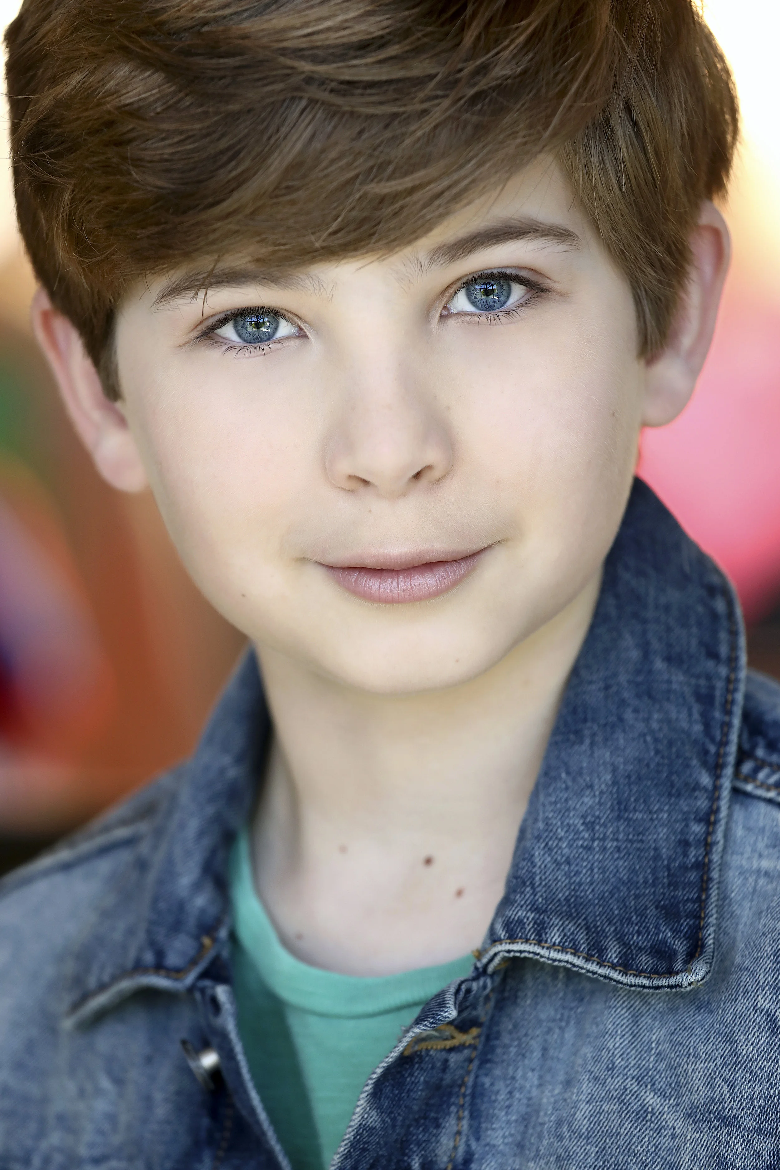 Close-up portrait of a young boy with light skin, blue eyes, short brown hair, wearing a denim jacket and teal shirt, smiling softly.