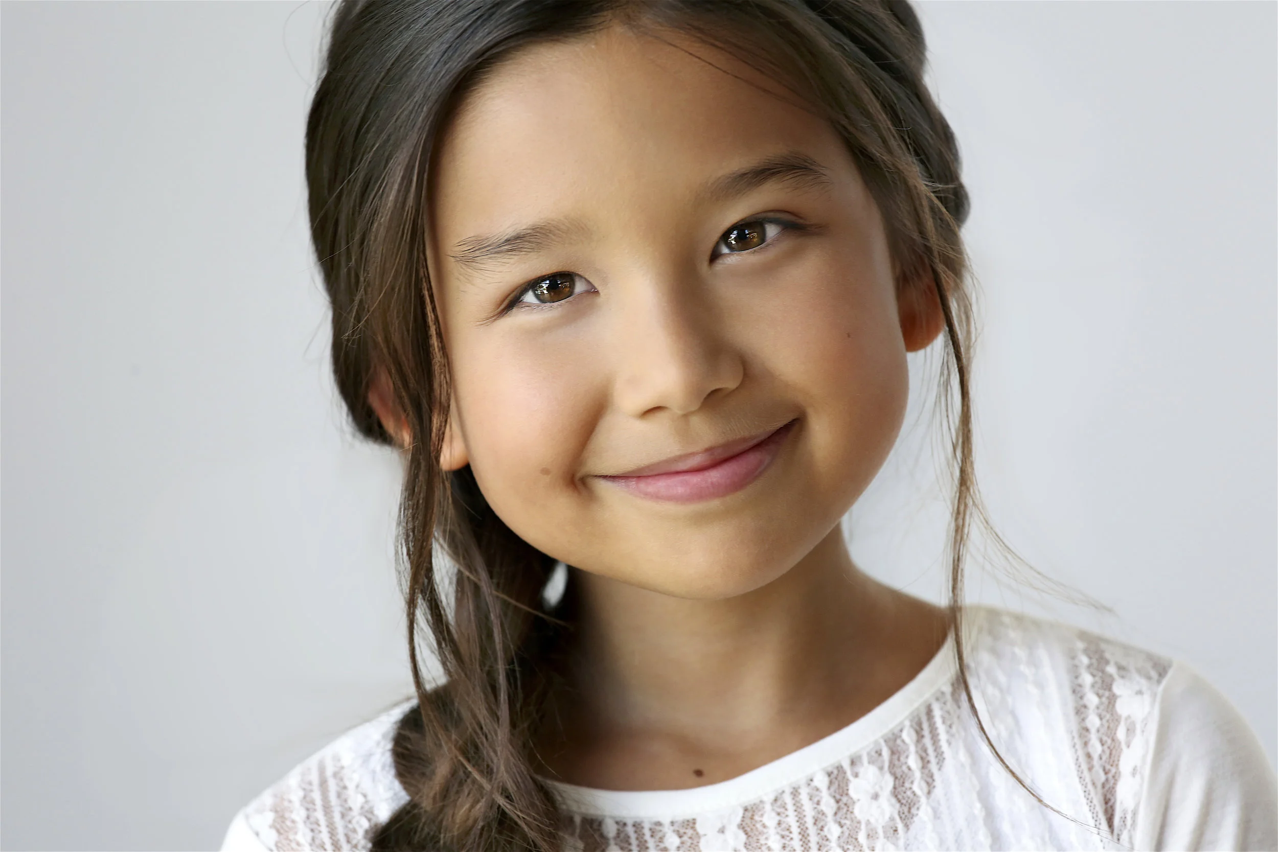 Close-up of a young girl with long dark hair smiling, wearing a white lace top, against a light gray background.