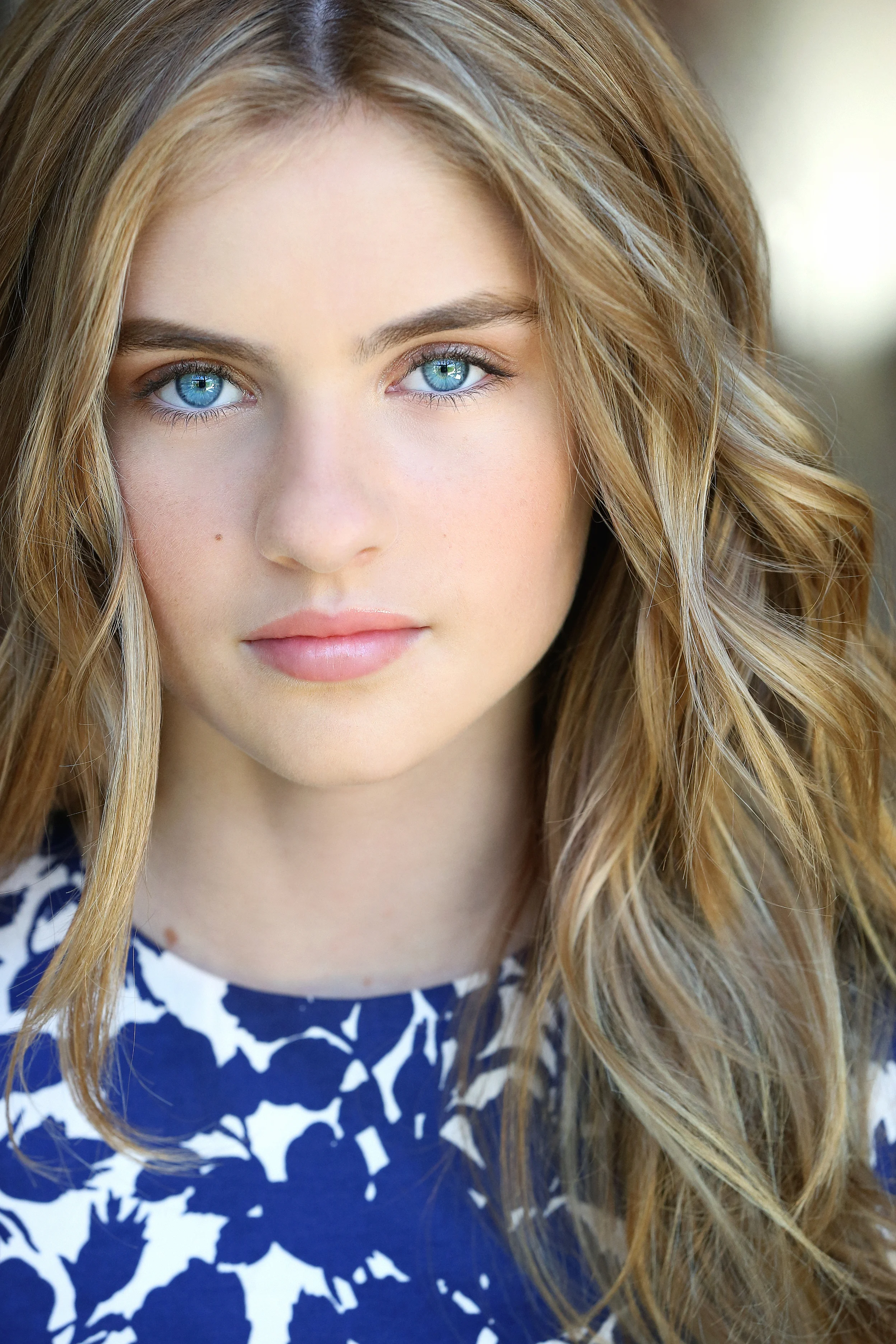 Close-up portrait of a young woman with blue eyes and long, wavy blonde hair, wearing a blue and white patterned top.