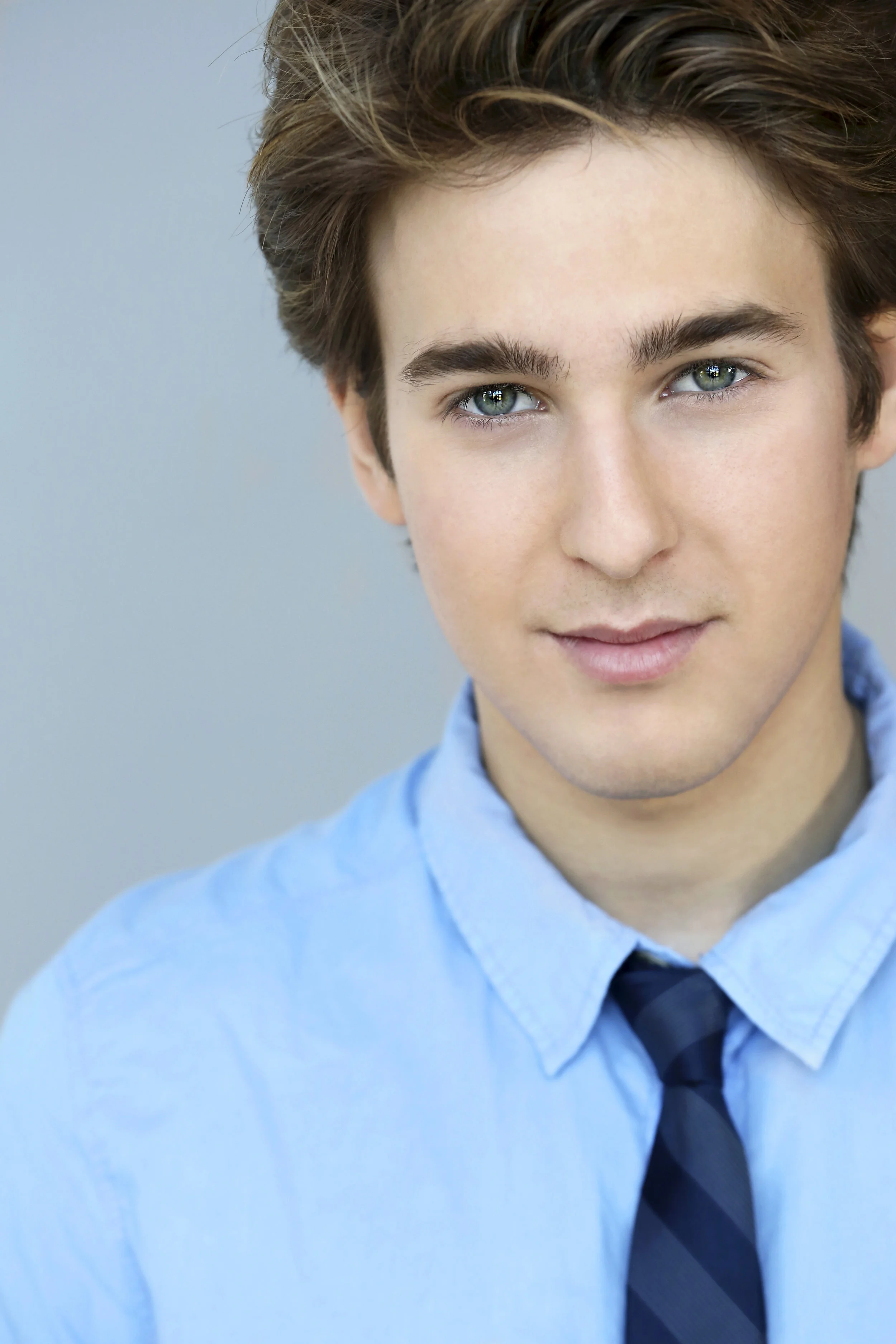 A young man with light brown hair and blue eyes wearing a light blue shirt and a dark striped tie, smiling softly against a plain background.