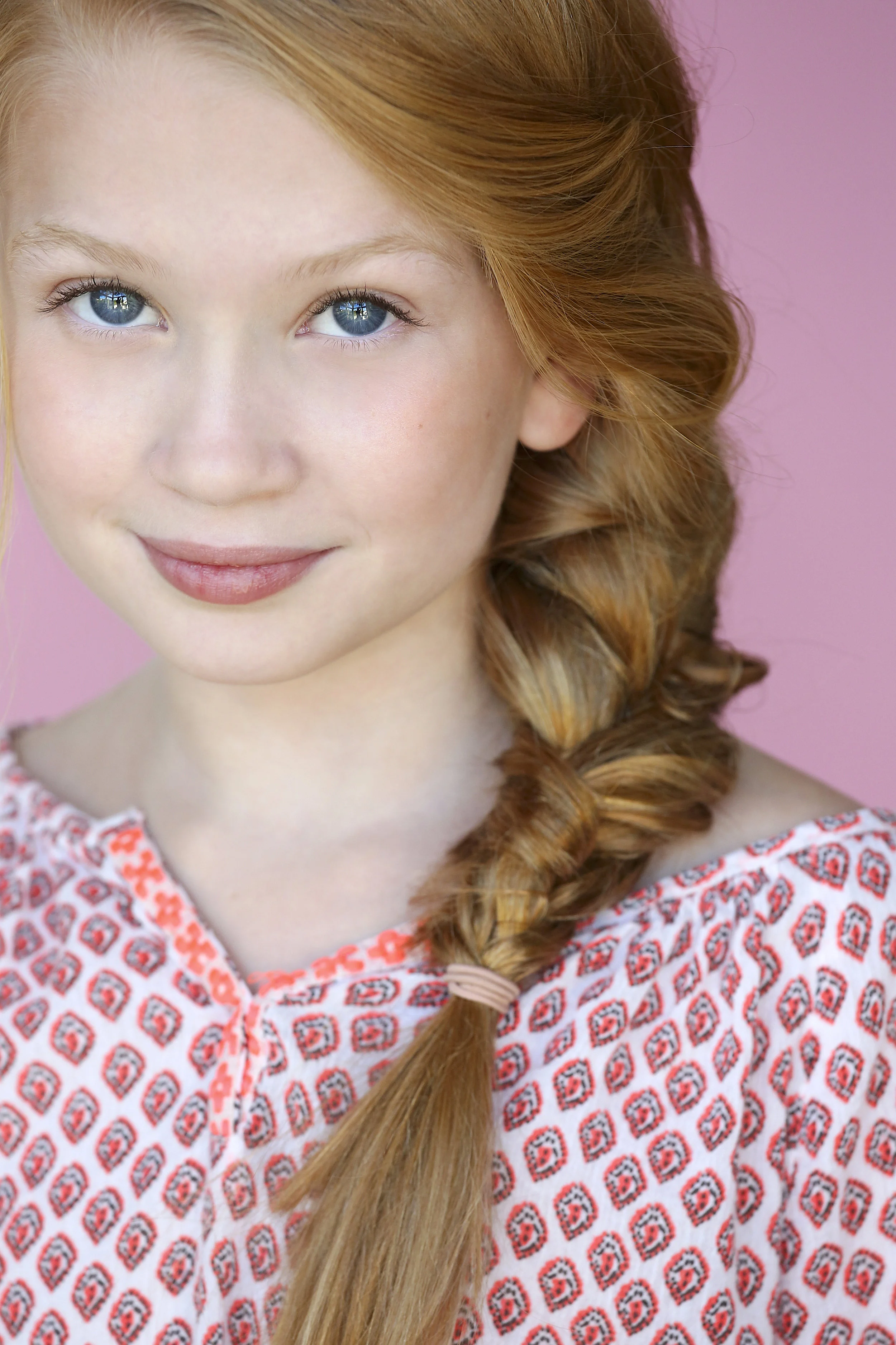 Close-up of a young girl with red hair styled in a loose braid, blue eyes, and a slight smile, wearing a patterned shirt, against a pink background.