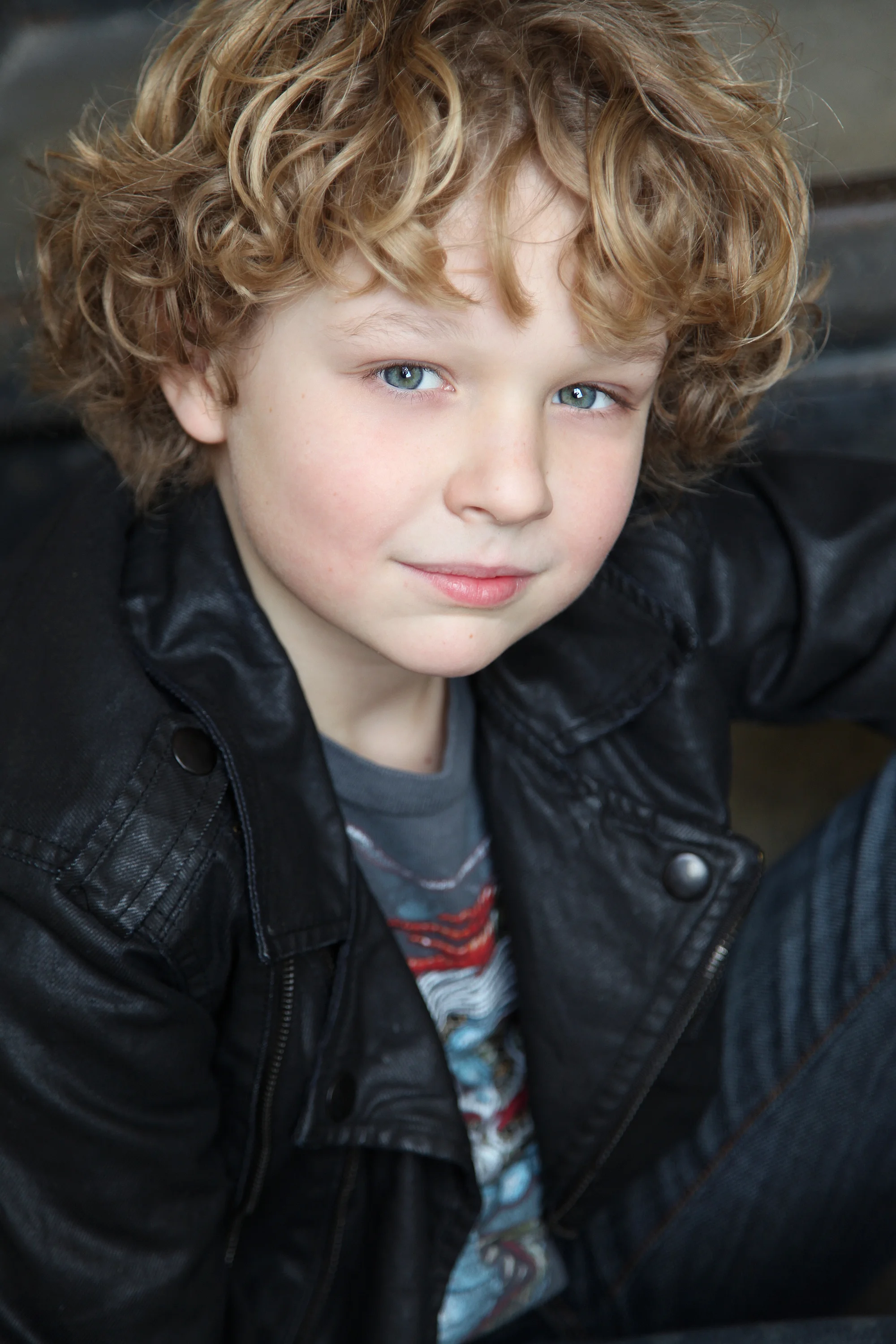 A young boy with curly red hair and blue eyes wearing a black leather jacket, looking at the camera with a slight smile.