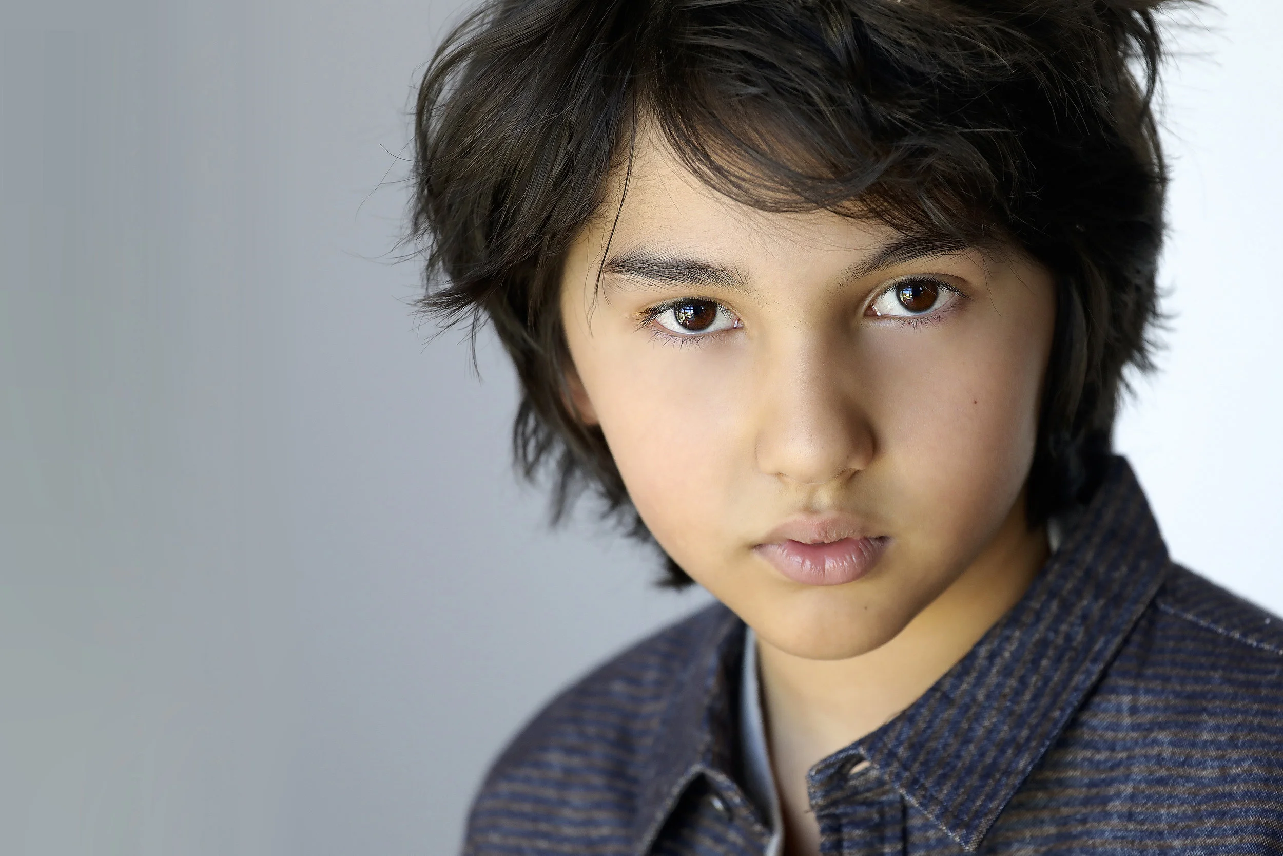 A young boy with dark, wavy hair and brown eyes looking directly at the camera, wearing a dark striped shirt, against a plain background.
