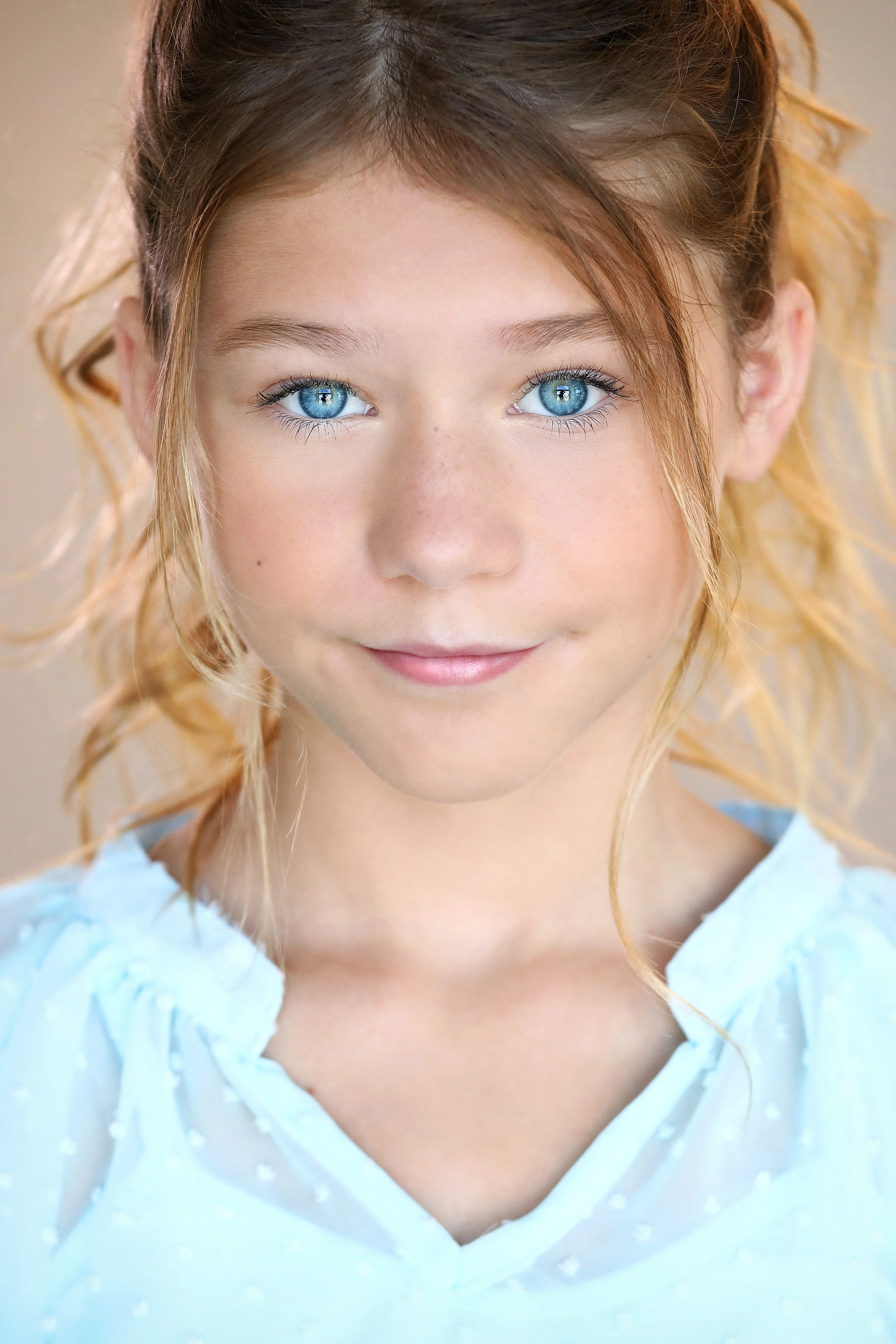 Close-up of a young girl with bright blue eyes, freckles, and blonde hair styled in loose curls, wearing a light blue top.