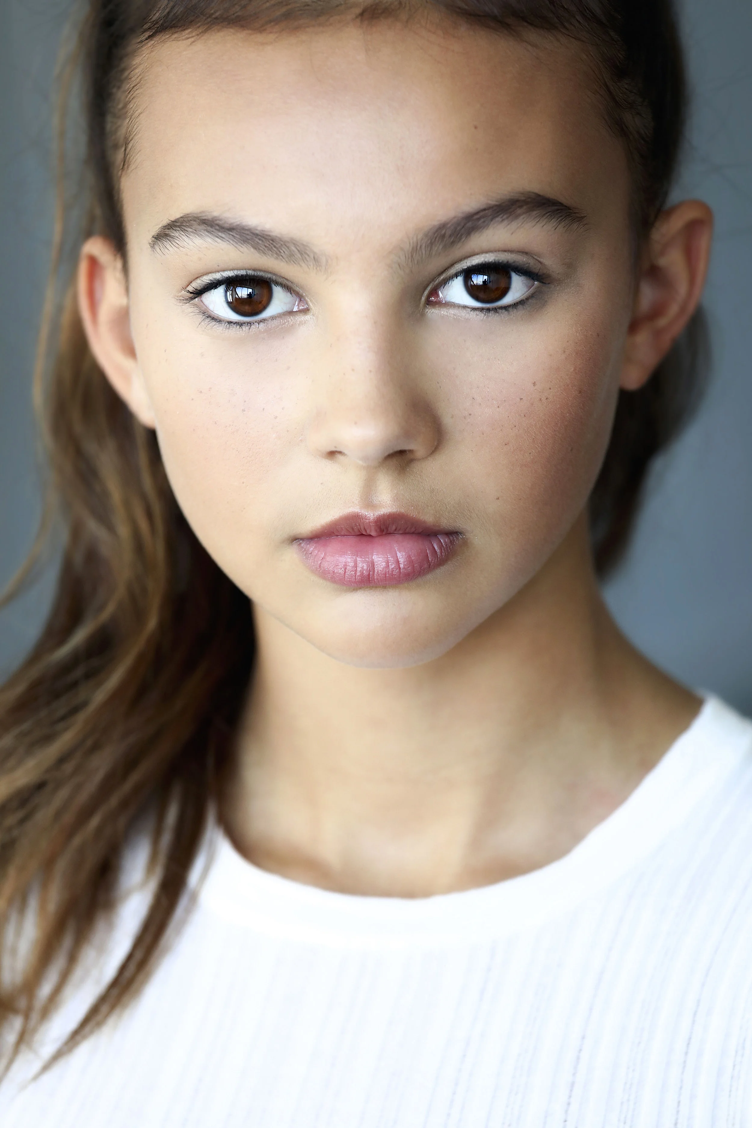 A close-up portrait of a young woman with brown eyes, light brown hair, and freckles, wearing a white top.
