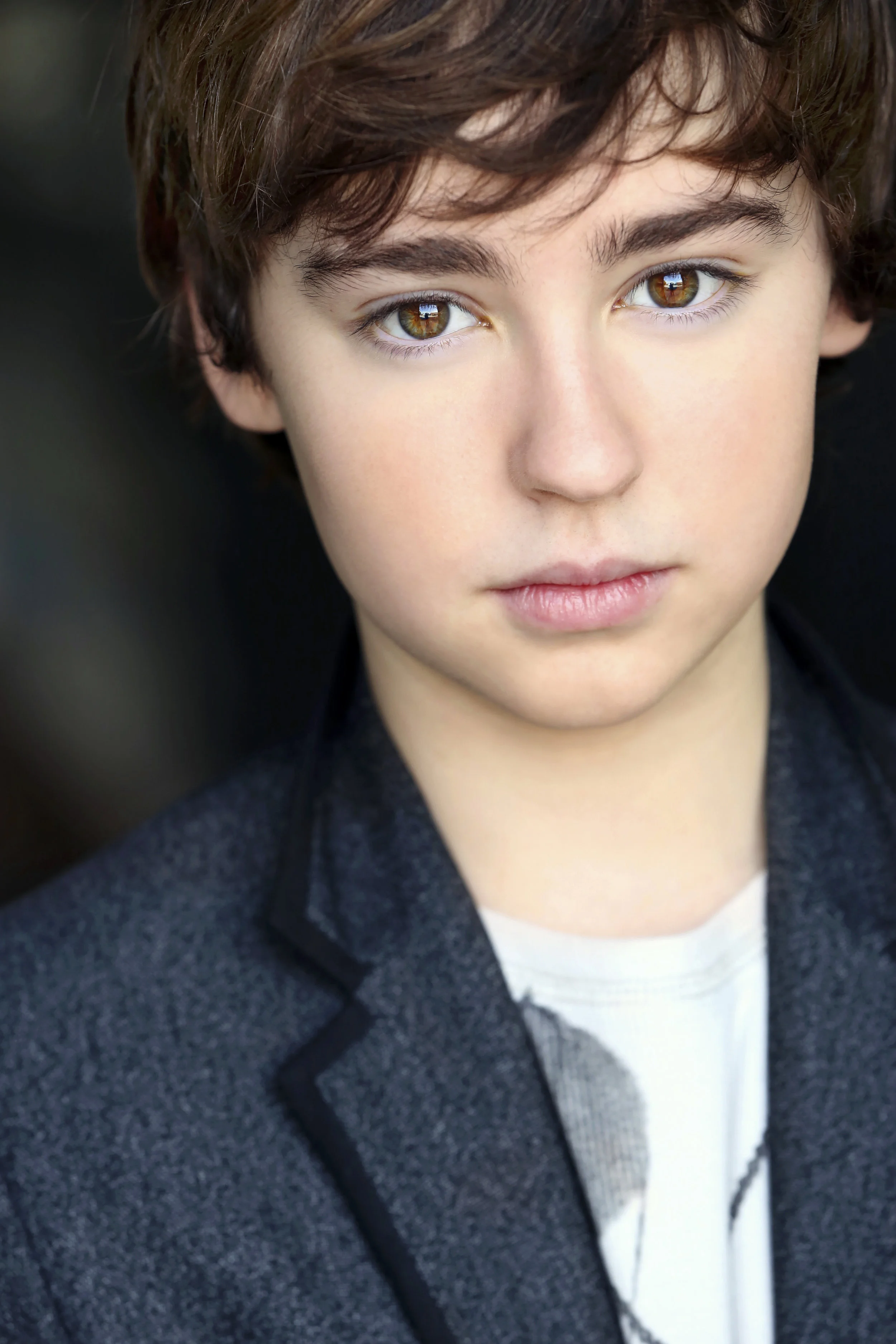 A close-up portrait of a young boy with brown hair and hazel eyes wearing a dark gray blazer and a white shirt.