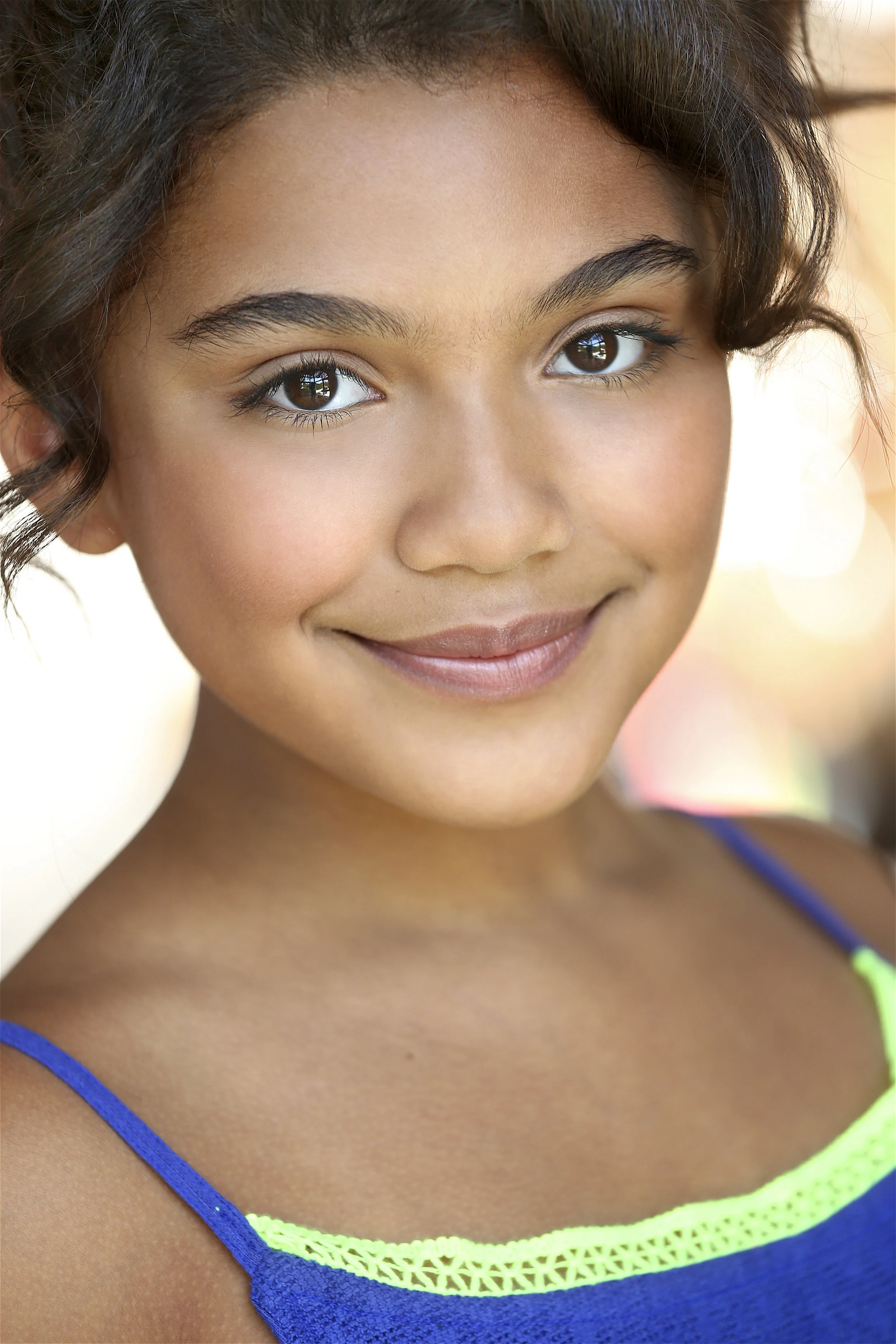 Close-up of a young girl with brown hair and brown eyes smiling, wearing a blue top with neon green trim.