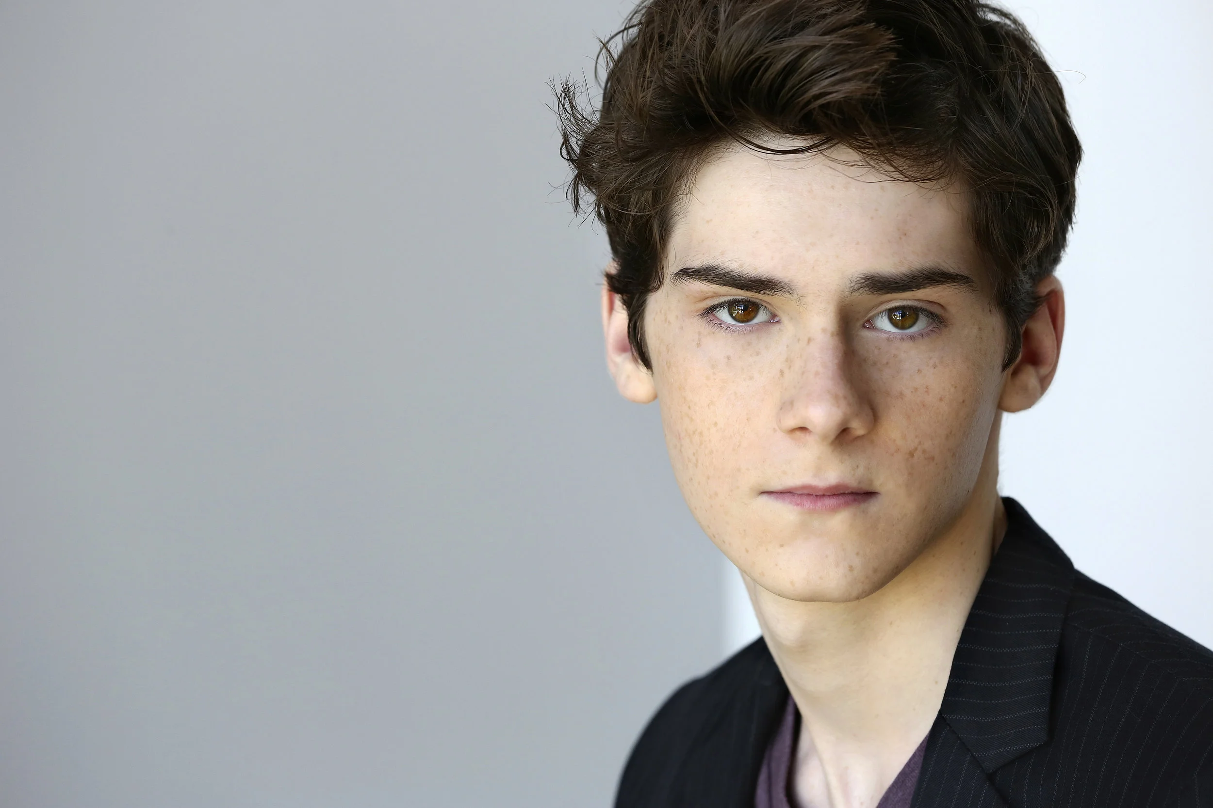 Close-up portrait of a young man with brown hair and hazel eyes, wearing a dark blazer, against a plain gray background.