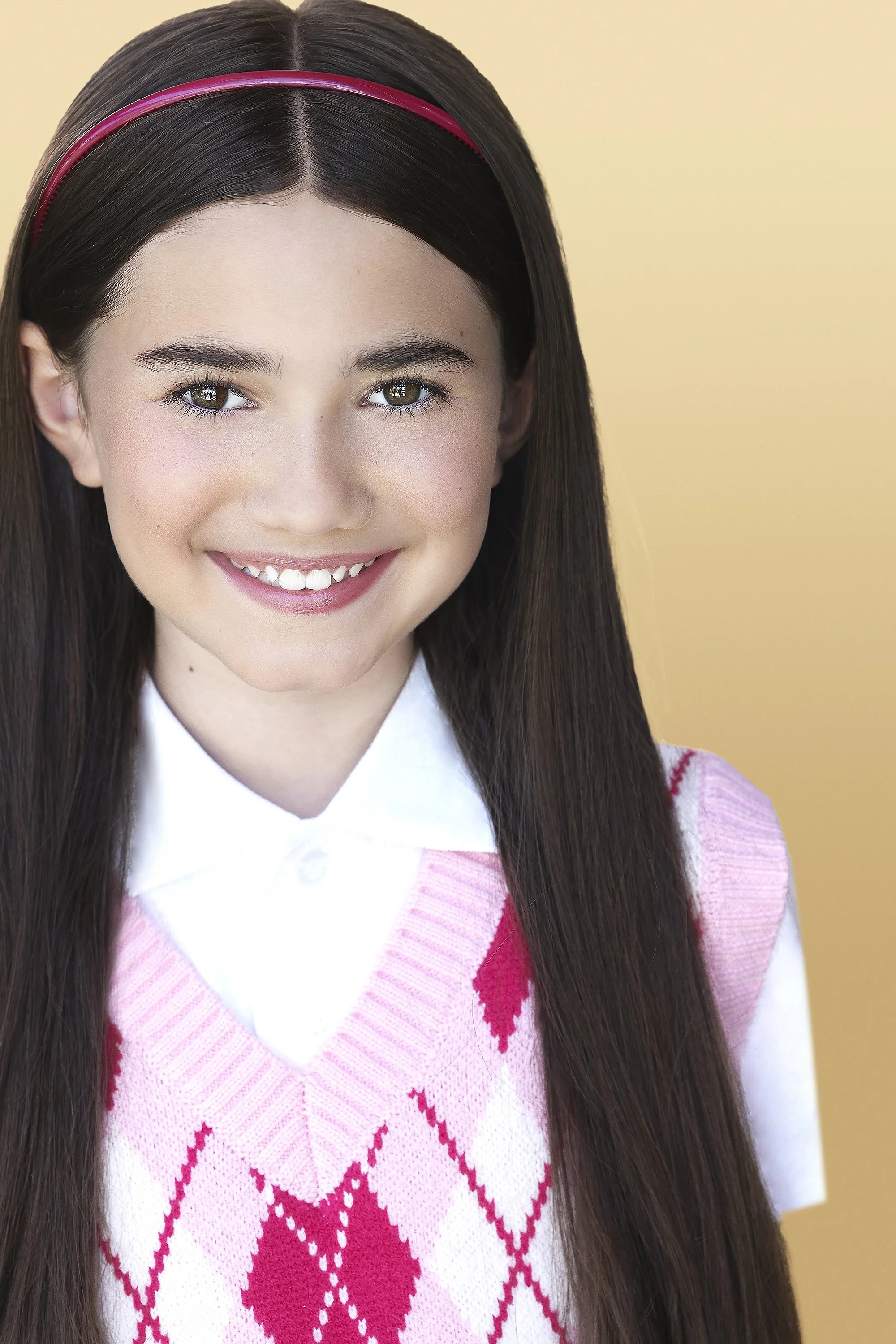 Close-up of a young girl with long brown hair, wearing a pink and white argyle sweater over a white collared shirt, smiling with a neutral yellow background.