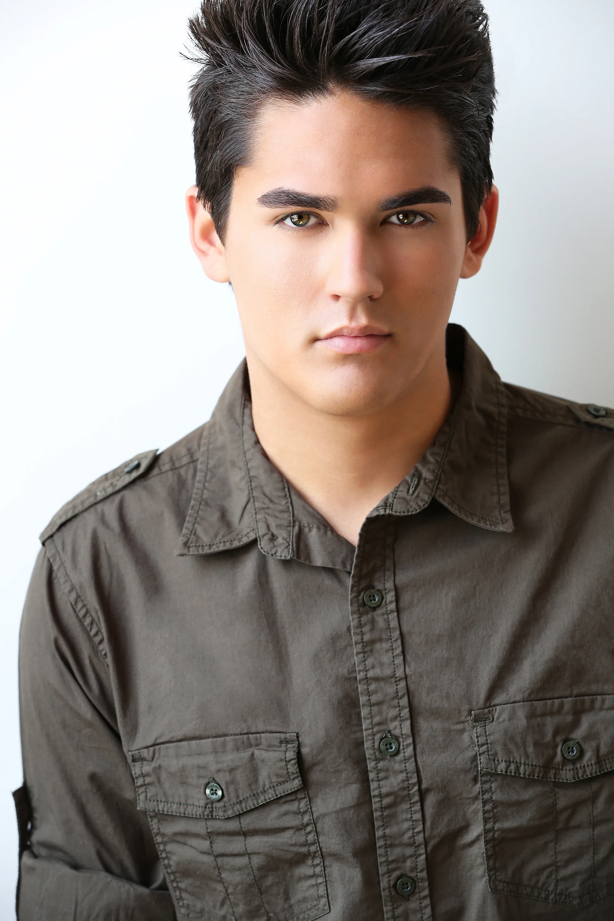 Portrait of a young man with dark, styled hair, wearing a gray button-up shirt with shoulder epaulets, against a plain background.