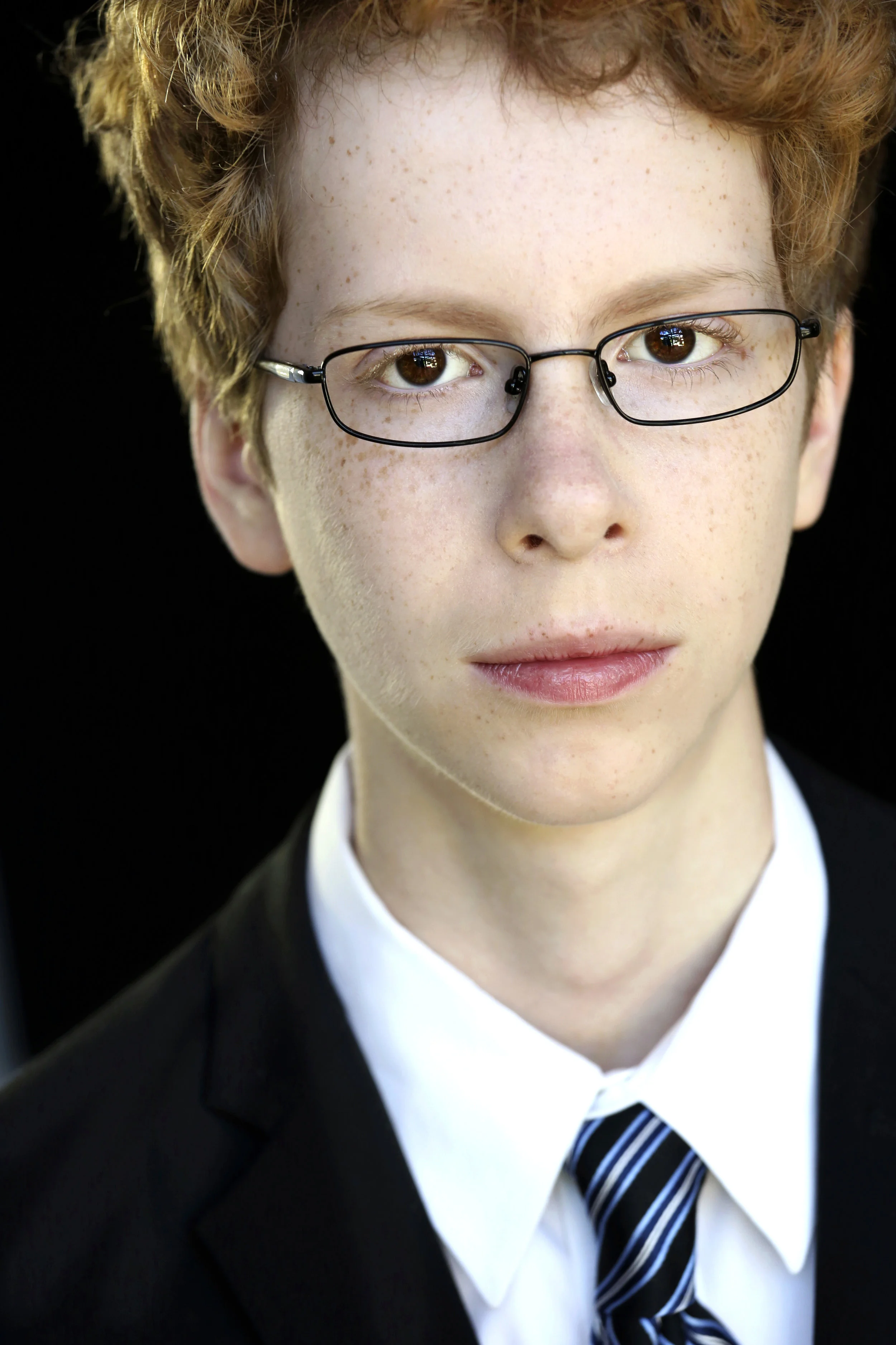Close-up portrait of a young boy with red hair, glasses, wearing a suit and tie, against a black background.