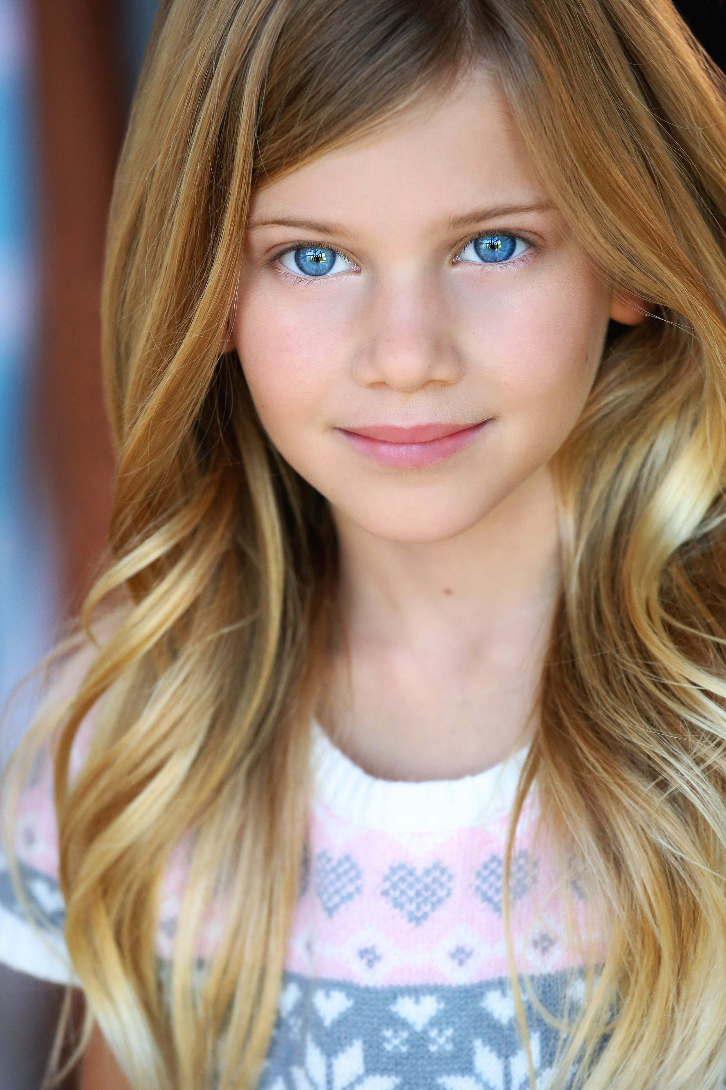 Close-up portrait of a young girl with long blonde hair and blue eyes, wearing a pink and gray sweater with a heart pattern.