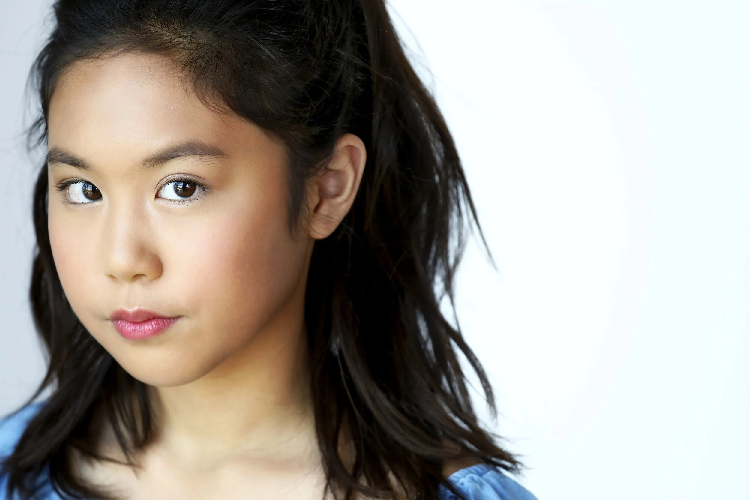 Close-up of a young girl with light brown skin and dark hair, looking directly at the camera against a plain light background.
