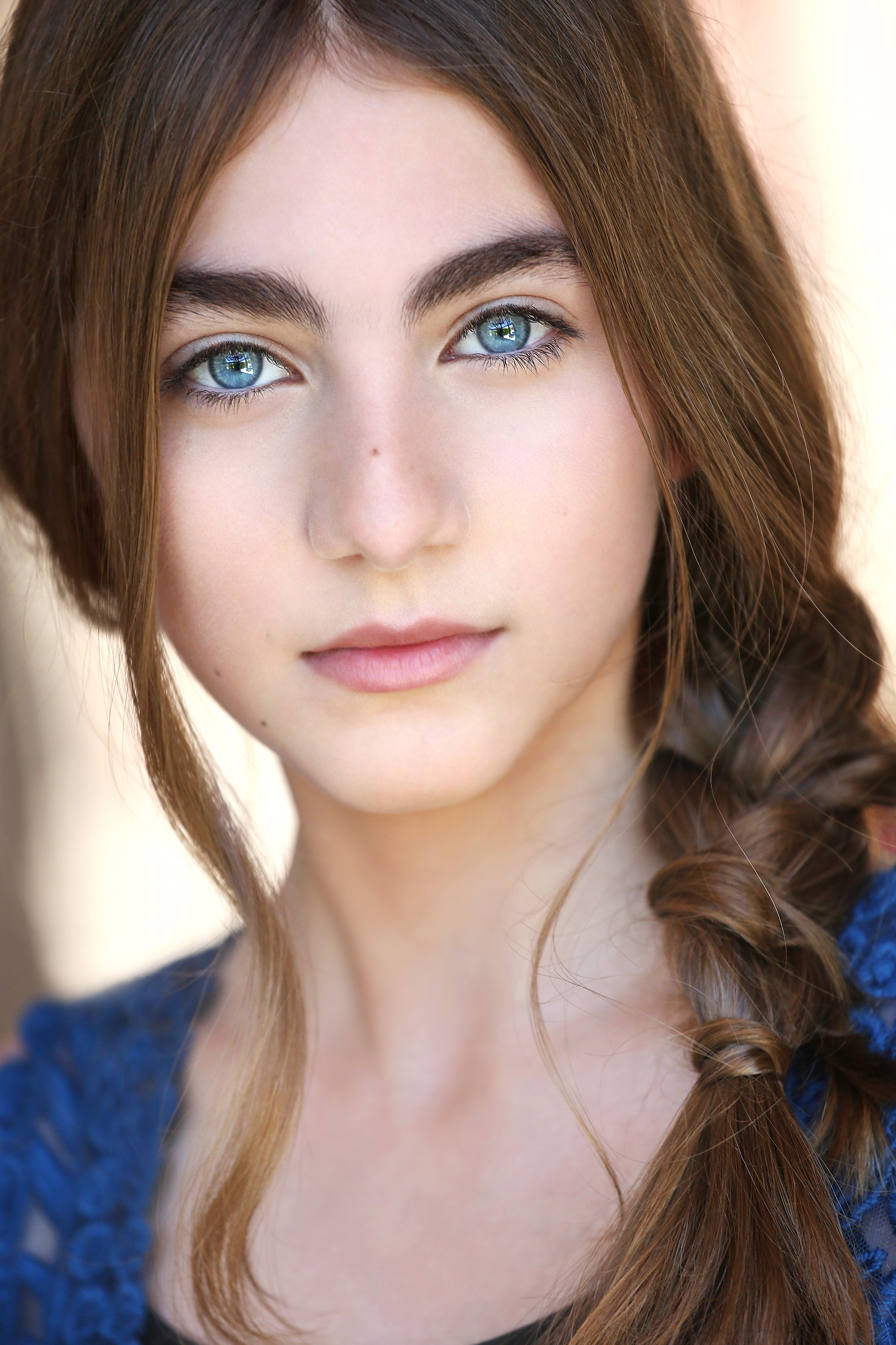 Close-up of a young woman with blue eyes, light makeup, long brown hair in a braid, wearing a blue top.