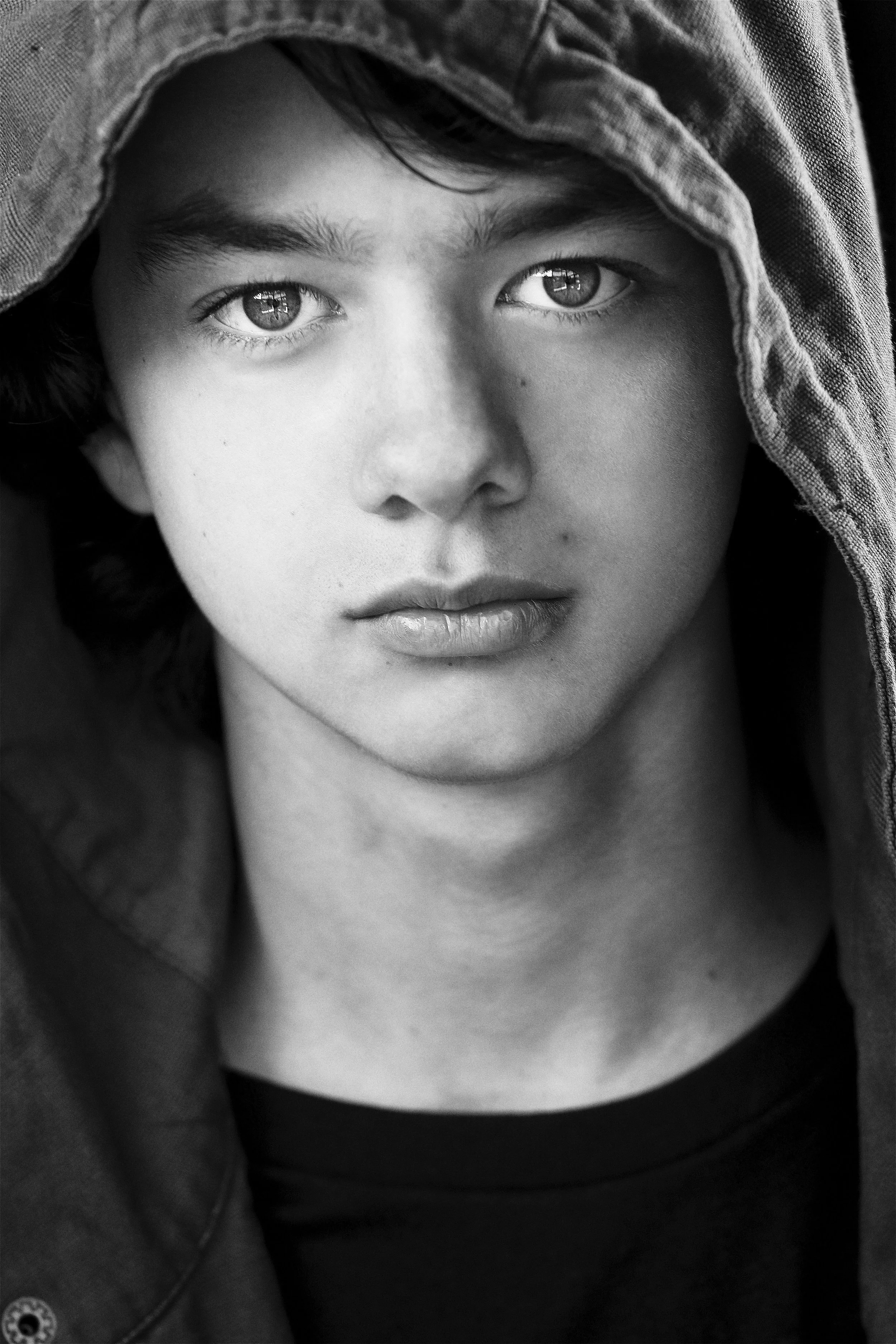 Black and white close-up portrait of a young person with light skin, wearing a hoodie and a black top, gazing directly at the camera.